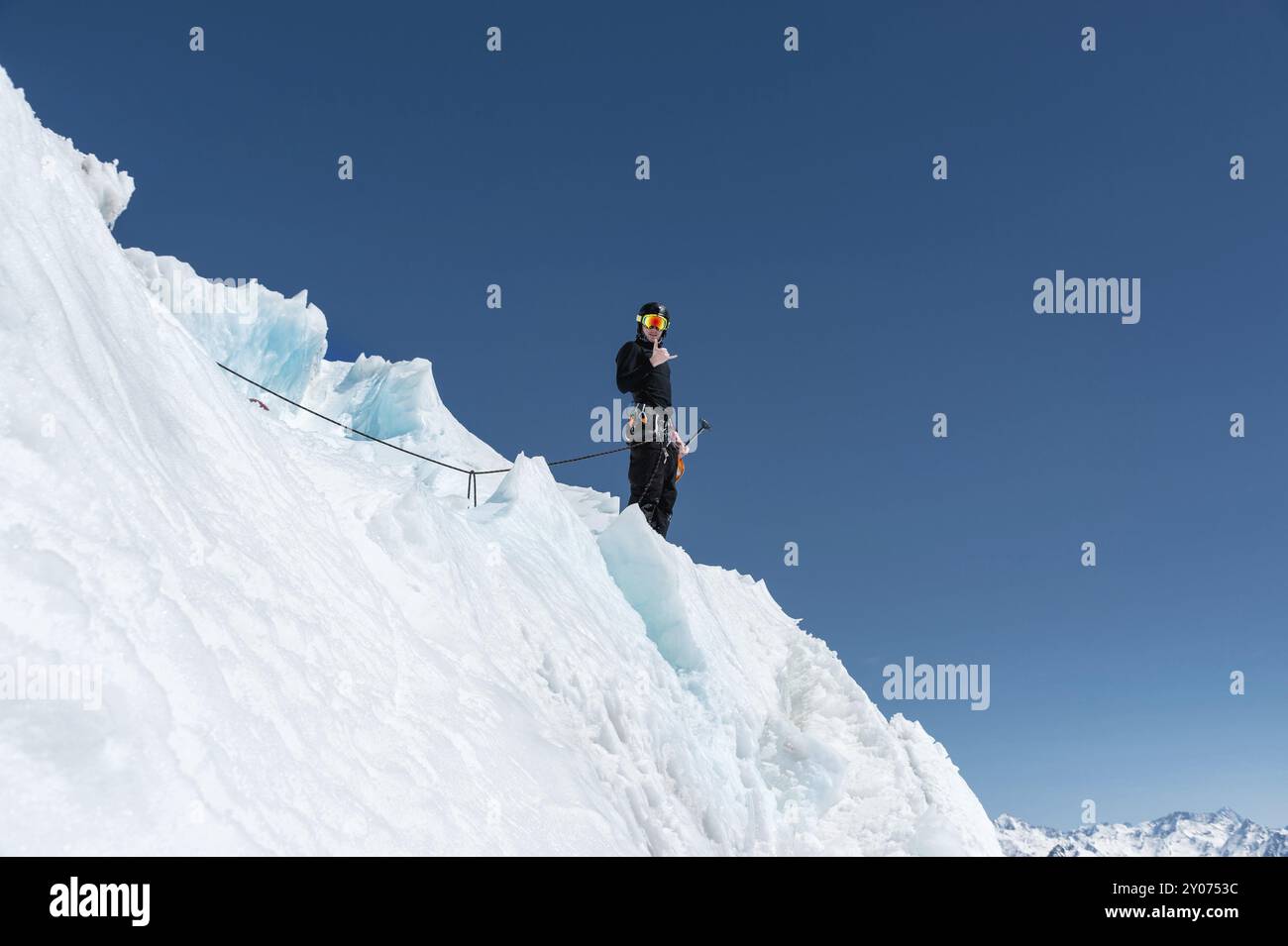 Un alpiniste se tient au bord d'un glacier avec une pelle à neige entre ses mains et montre le geste de Shak contre le ciel bleu Banque D'Images