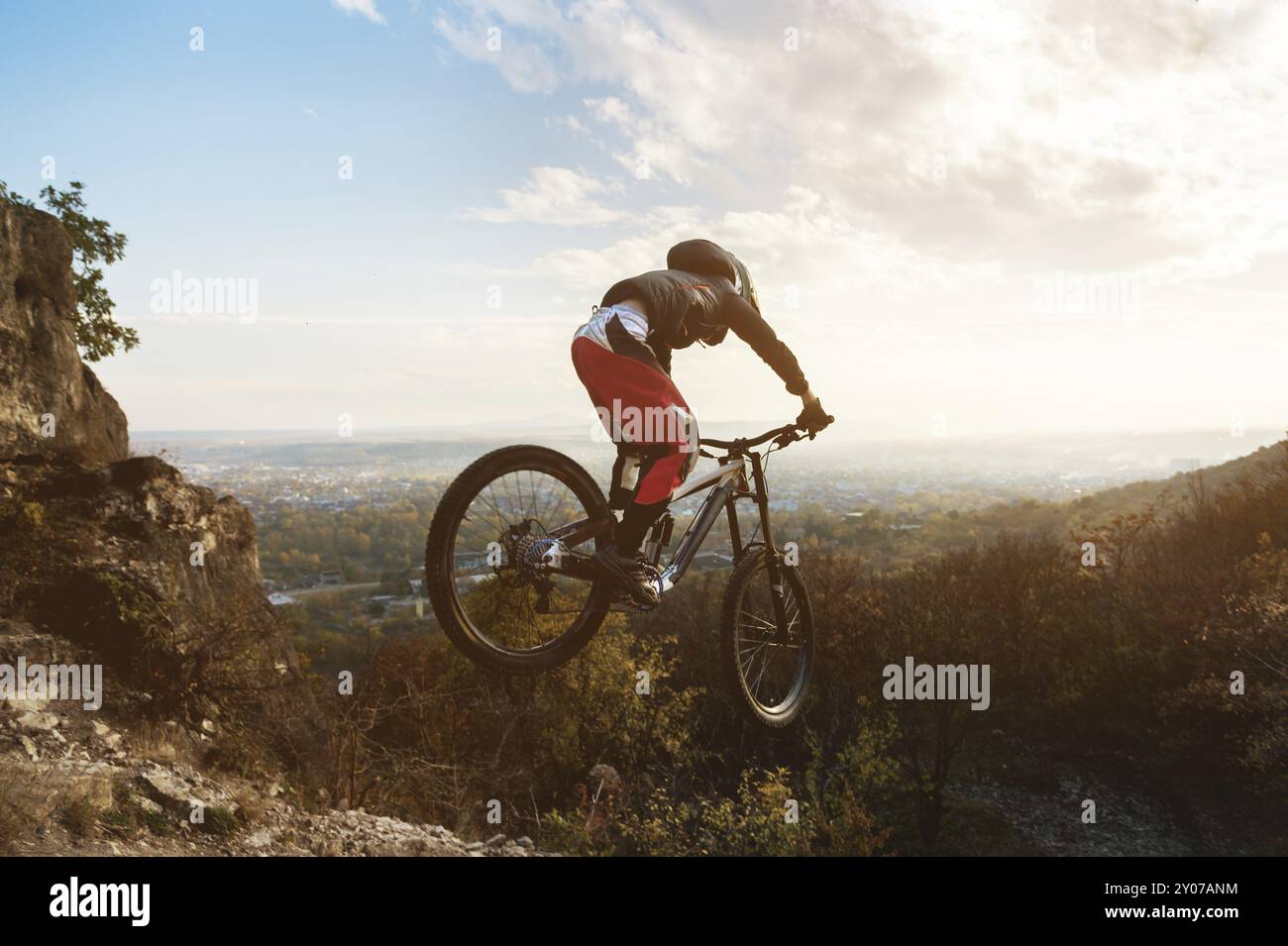 Un jeune pilote au volant de son VTT saute de hautes falaises sur fond de paysage urbain au coucher du soleil. sentier de montagne descendant en t Banque D'Images