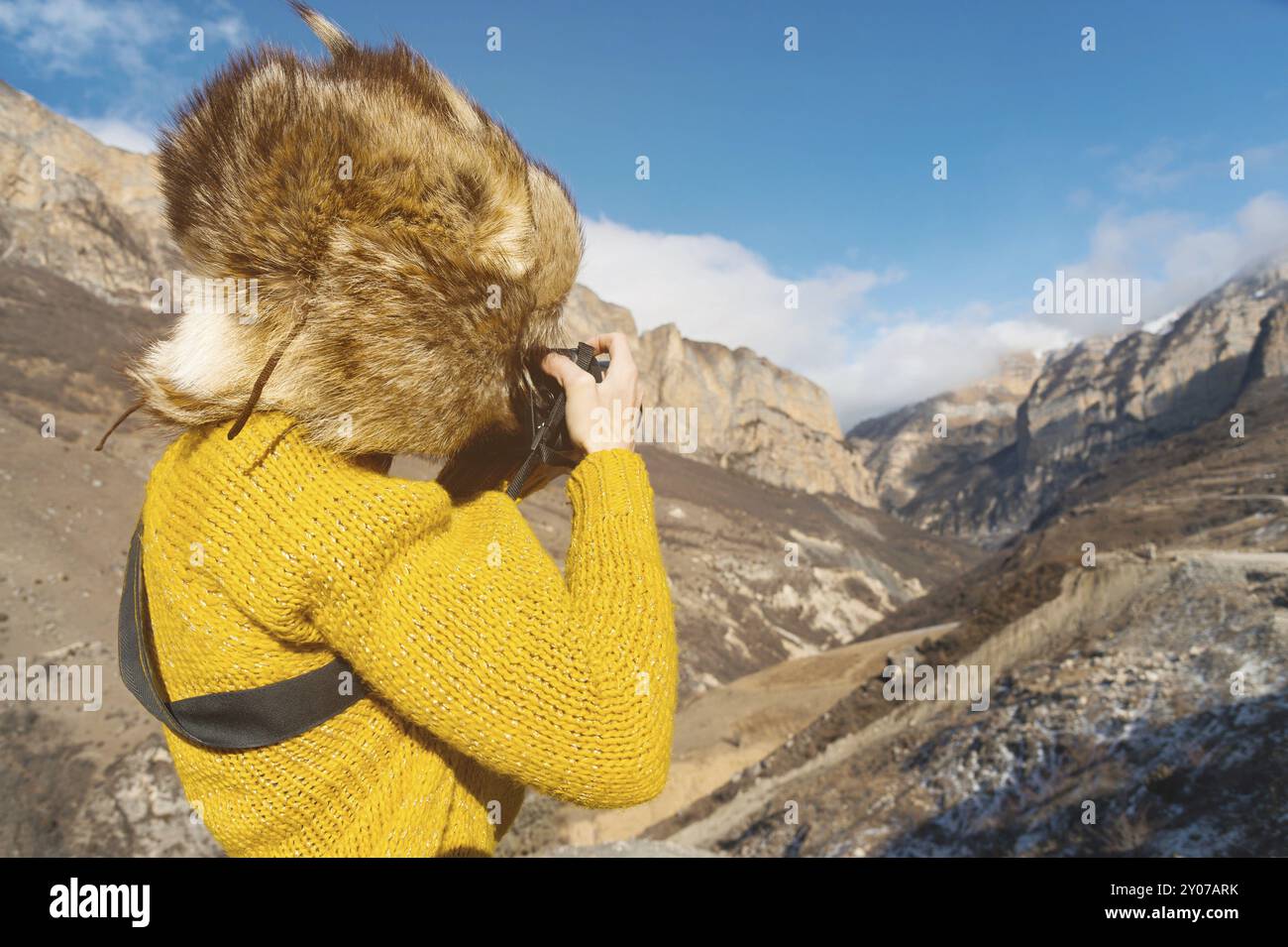 Jeune photographe touristique dans un chapeau de fourrure et un pull jaune dans les montagnes prend des photos sur son appareil photo numérique. Le concept de photographie dans le voyage Banque D'Images