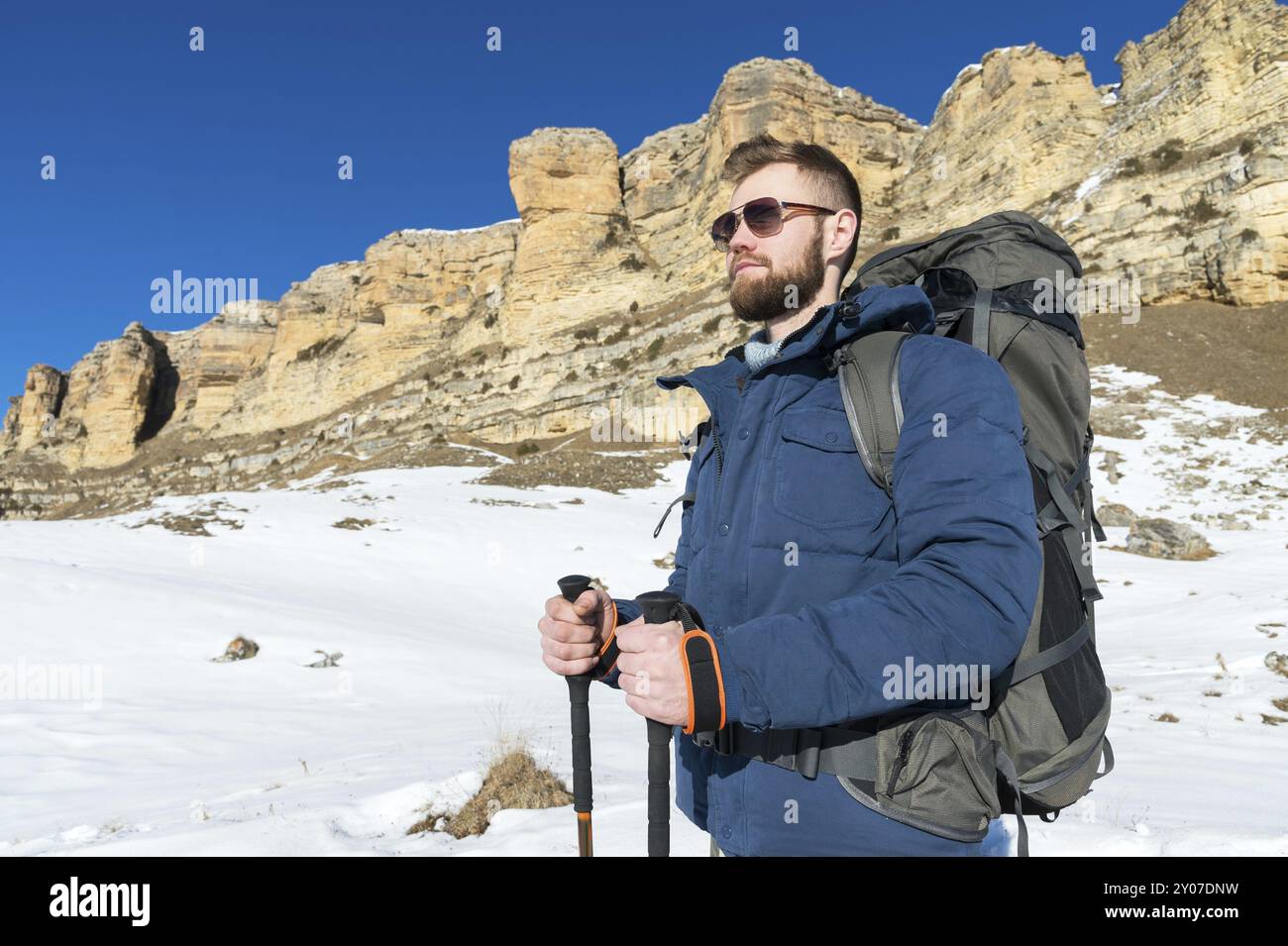 Portrait Un hipster barbu avec un sac à dos et dans des lunettes de soleil avec un grand sac à dos sur ses épaules se dresse avec des bâtons pour la marche nordique sur le dos Banque D'Images