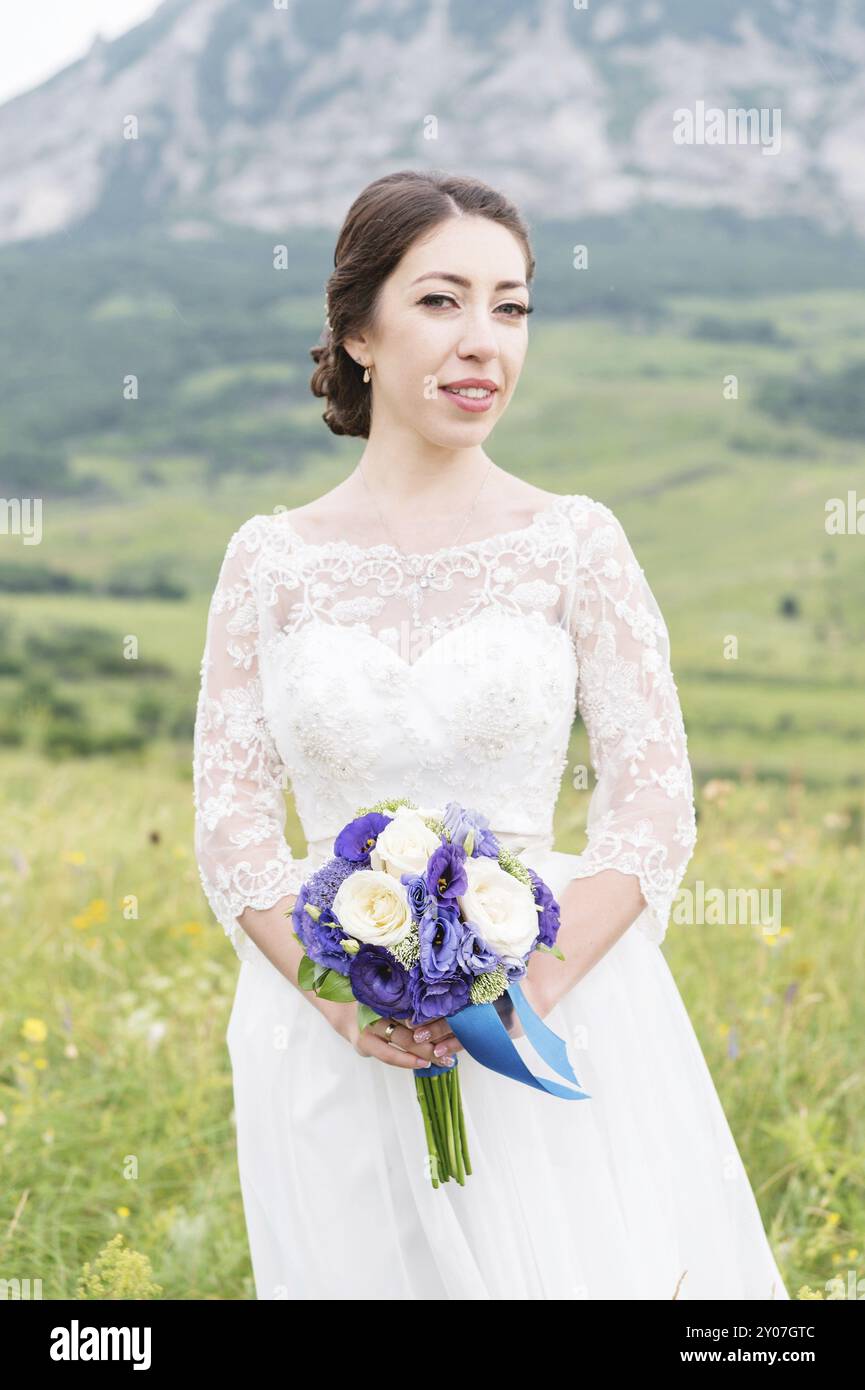 Une belle mariée caucasienne avec un bouquet de fleurs de mariage dans ses mains se tient sur le fond des montagnes du Caucase et des champs verts Banque D'Images