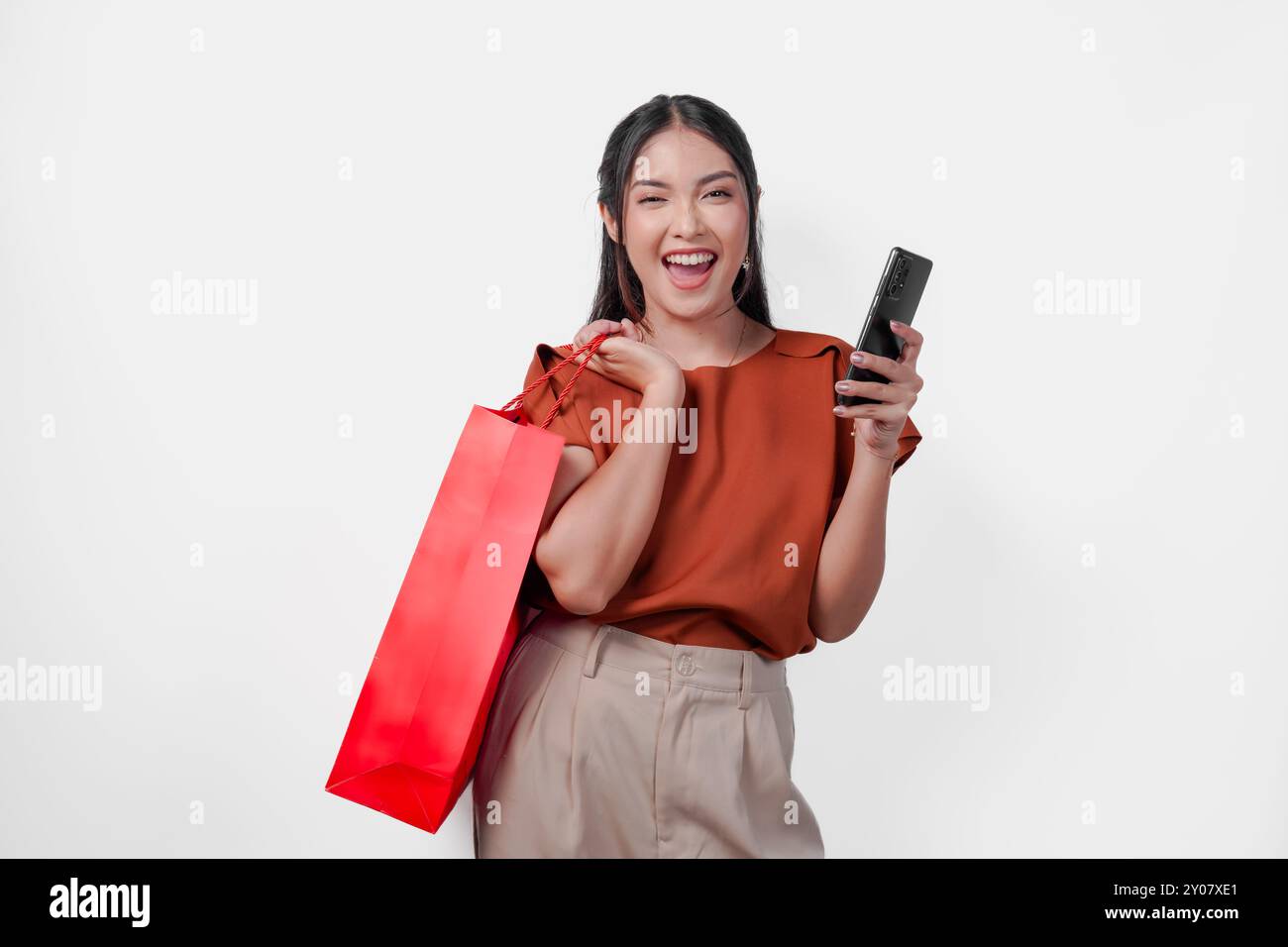 Heureuse jeune femme asiatique tient un sac à provisions rouge tout en tenant un téléphone portable. Concept de bonheur et d'excitation, éventuellement lié à un shopping Banque D'Images