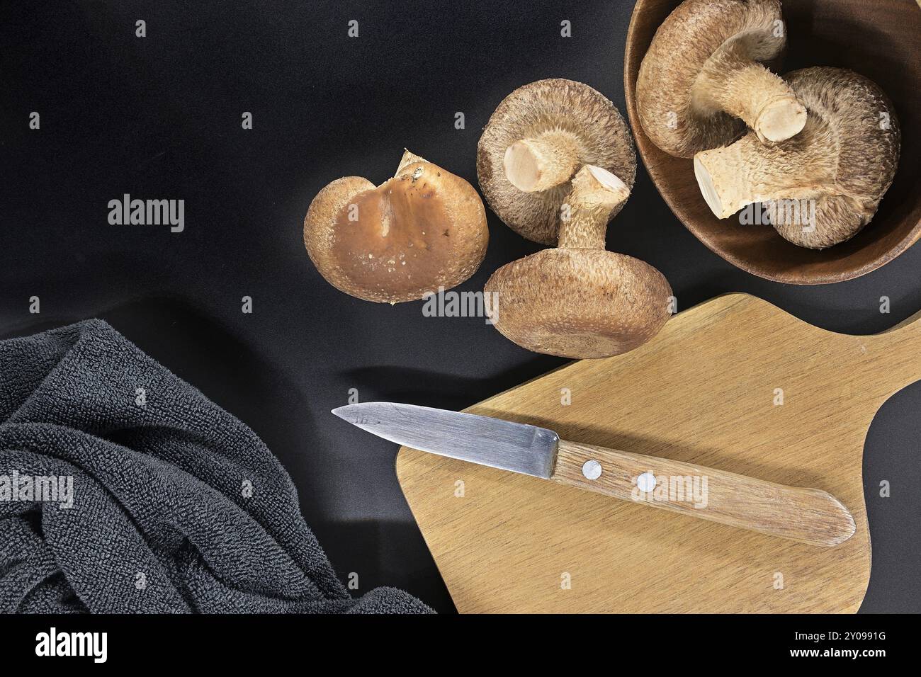 Bol contenant des champignons shiitake frais, planche à découper en bois avec couteau dessus et serviette sombre disposée sur fond noir Banque D'Images
