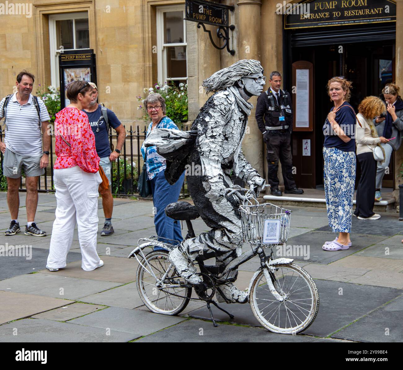 Un artiste vêtu d'un costume unique et accrocheur monte sur un vélo blanc devant le bâtiment historique de la salle des pompes, en observant les passants. Banque D'Images