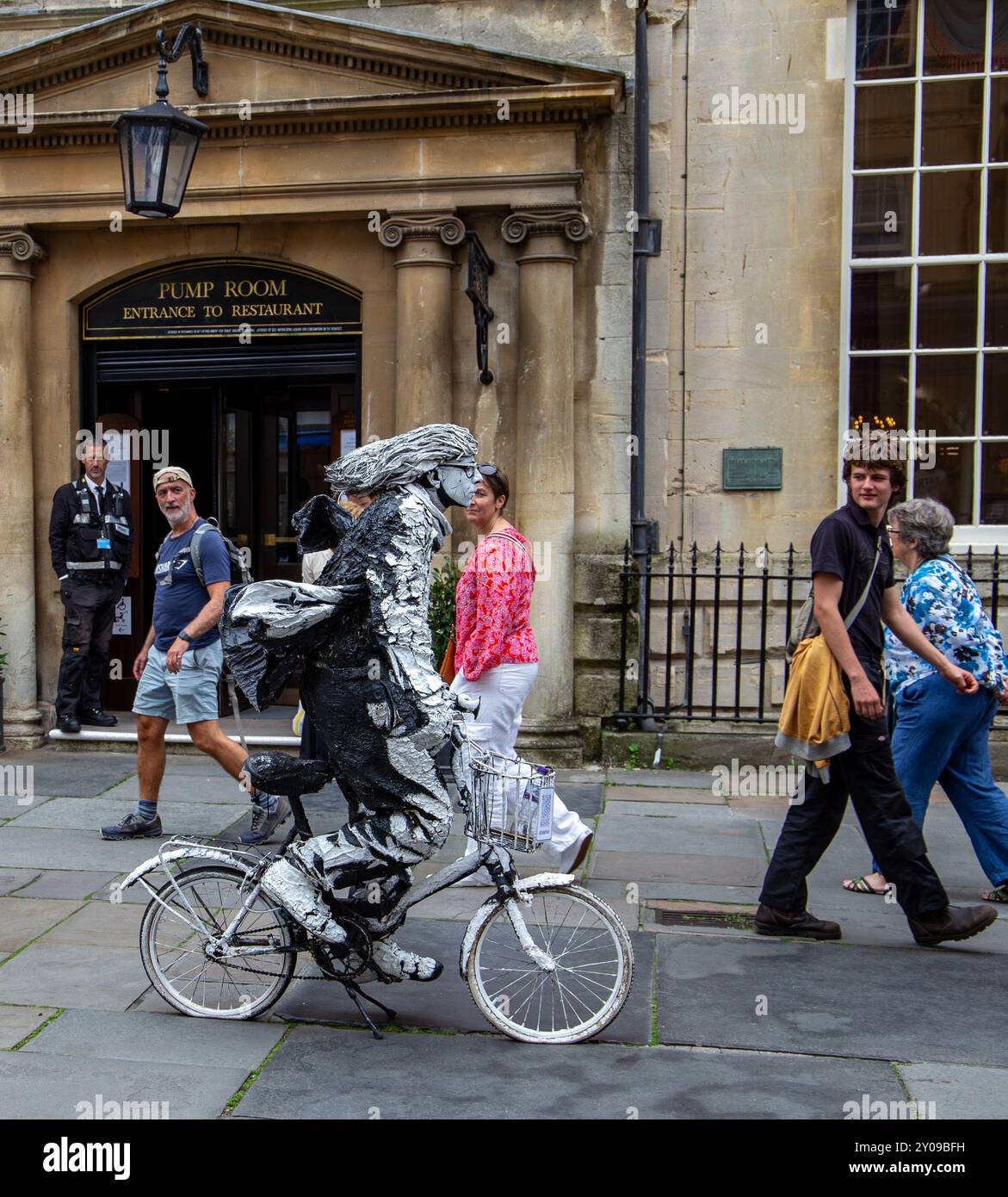 Un artiste vêtu d'un costume unique et accrocheur monte sur un vélo blanc devant le bâtiment historique de la salle des pompes, en observant les passants. Banque D'Images