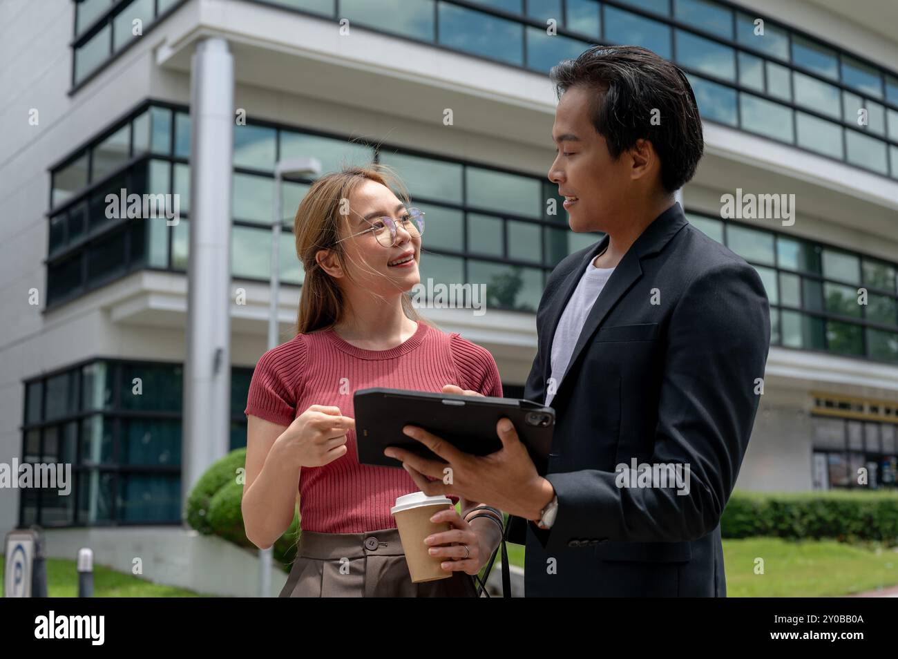 Une belle assistante ou secrétaire asiatique positive discute du travail avec son patron tout en marchant à l'extérieur, avec un bâtiment moderne dans le dos Banque D'Images