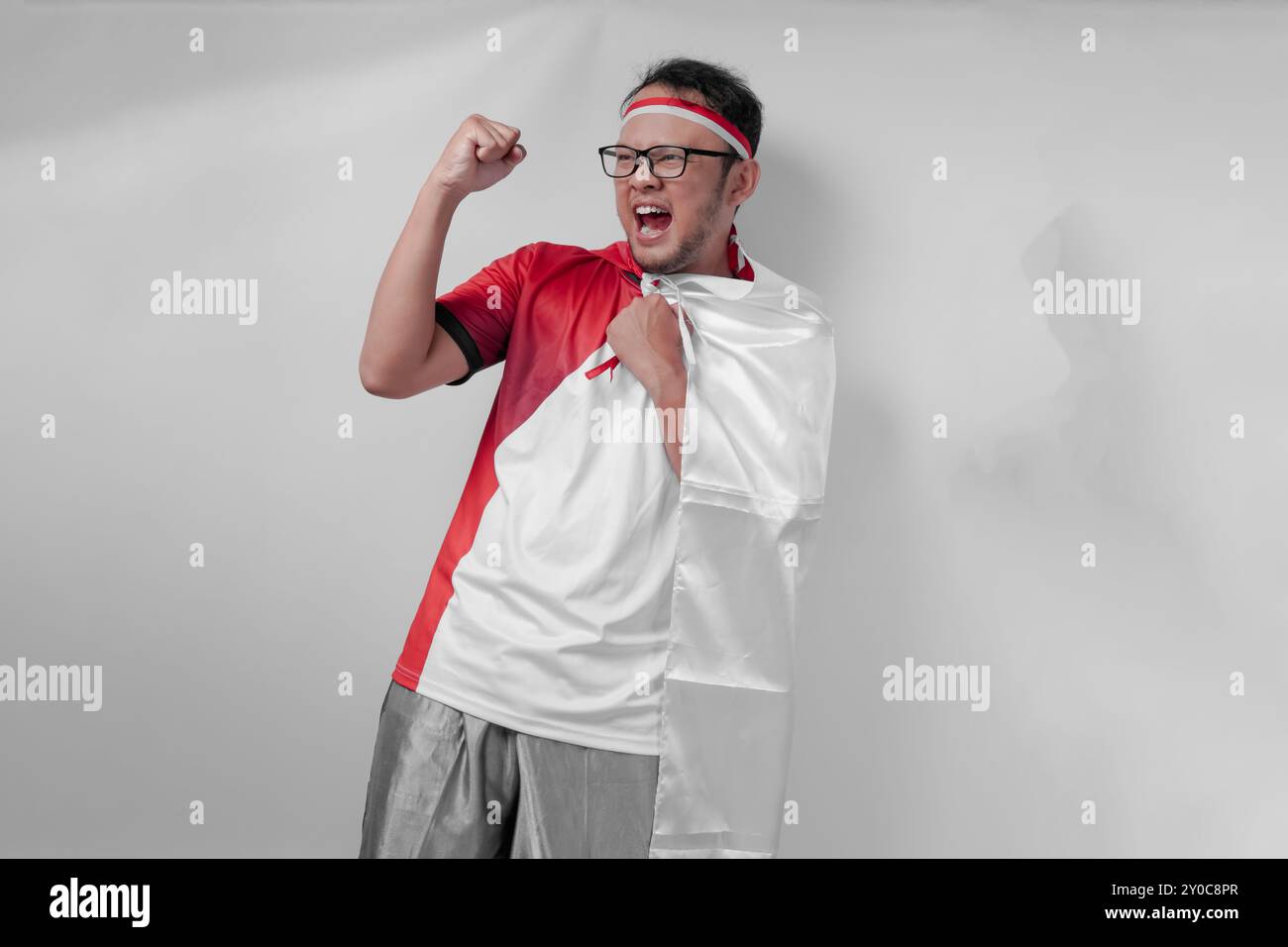 Jeune homme asiatique énergique portant un maillot avec drapeau drapé sur l'épaule levant la main dans les airs avec une expression heureuse. Jour de l'indépendance de l'Indonésie et Banque D'Images