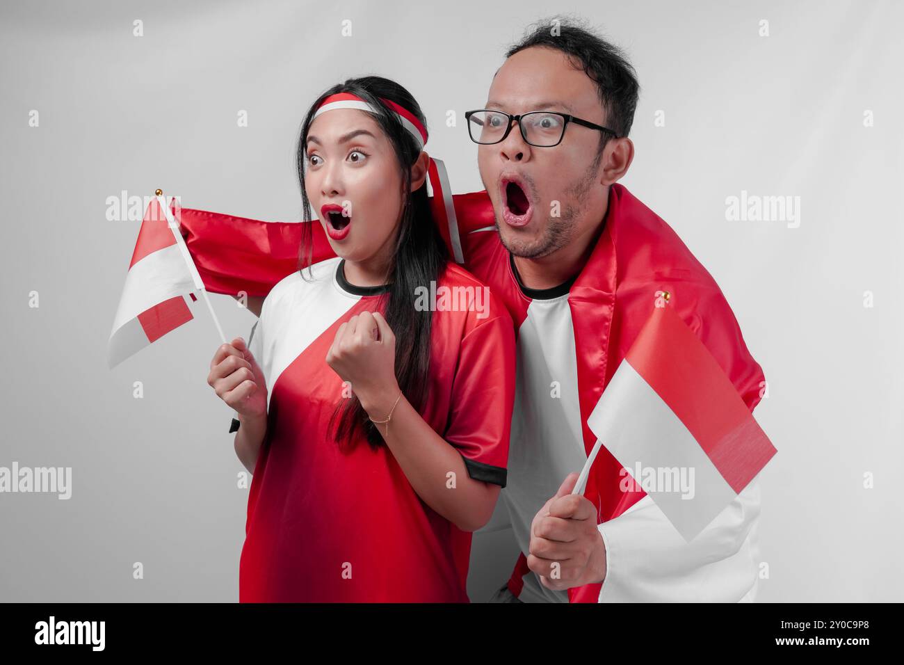 Couple indonésien choqué tenant des drapeaux nationaux tout en regardant la compétition sportive ensemble. Jour de l'indépendance de l'Indonésie et concept sportif. Banque D'Images