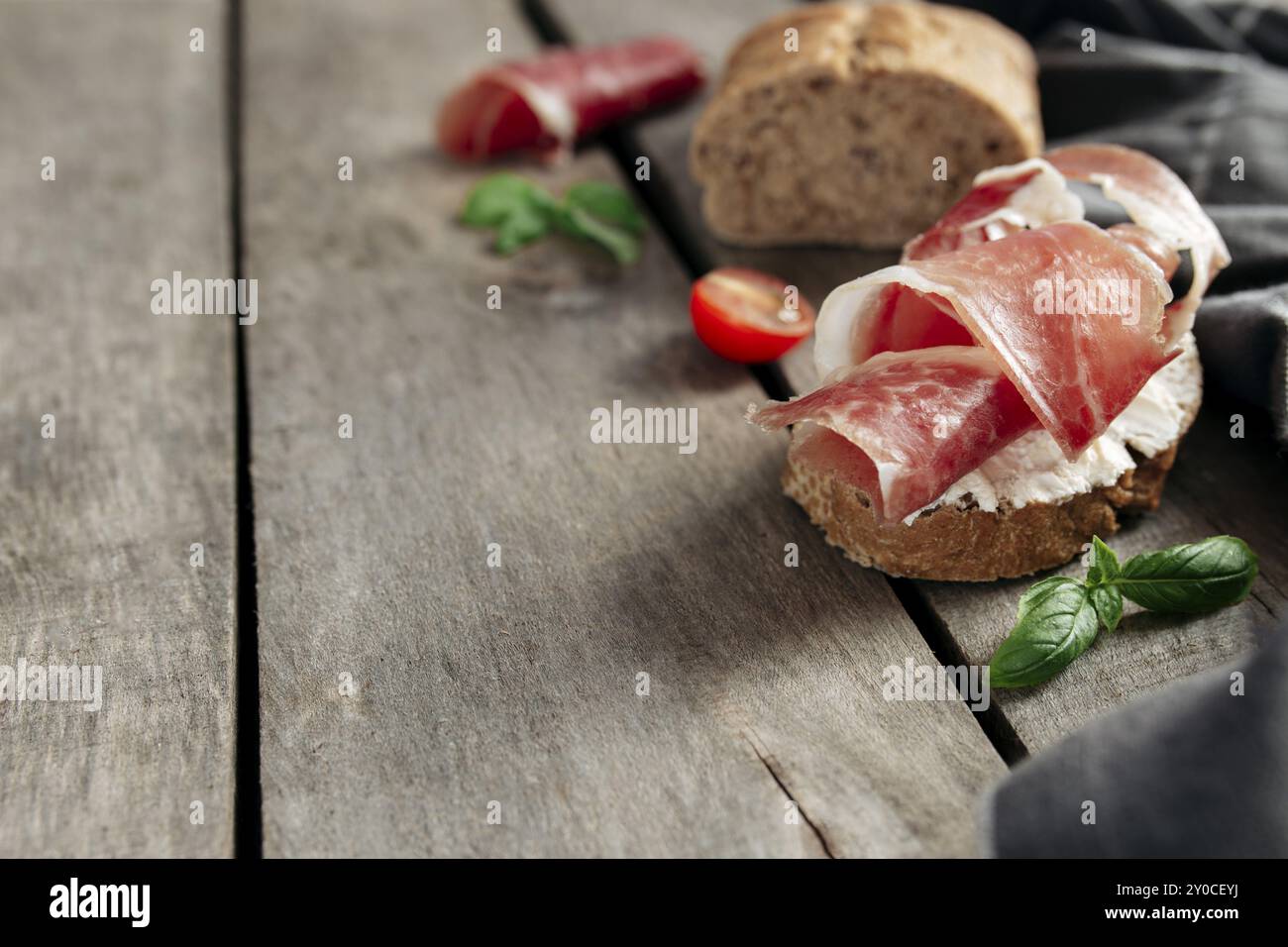 Concept de cuisine italienne. Pain croustillant grillé au fromage à la crème et émincé de jambon, tomates cerises, branches de feuilles de basilic, serviette de cuisine sur la ba en bois Banque D'Images
