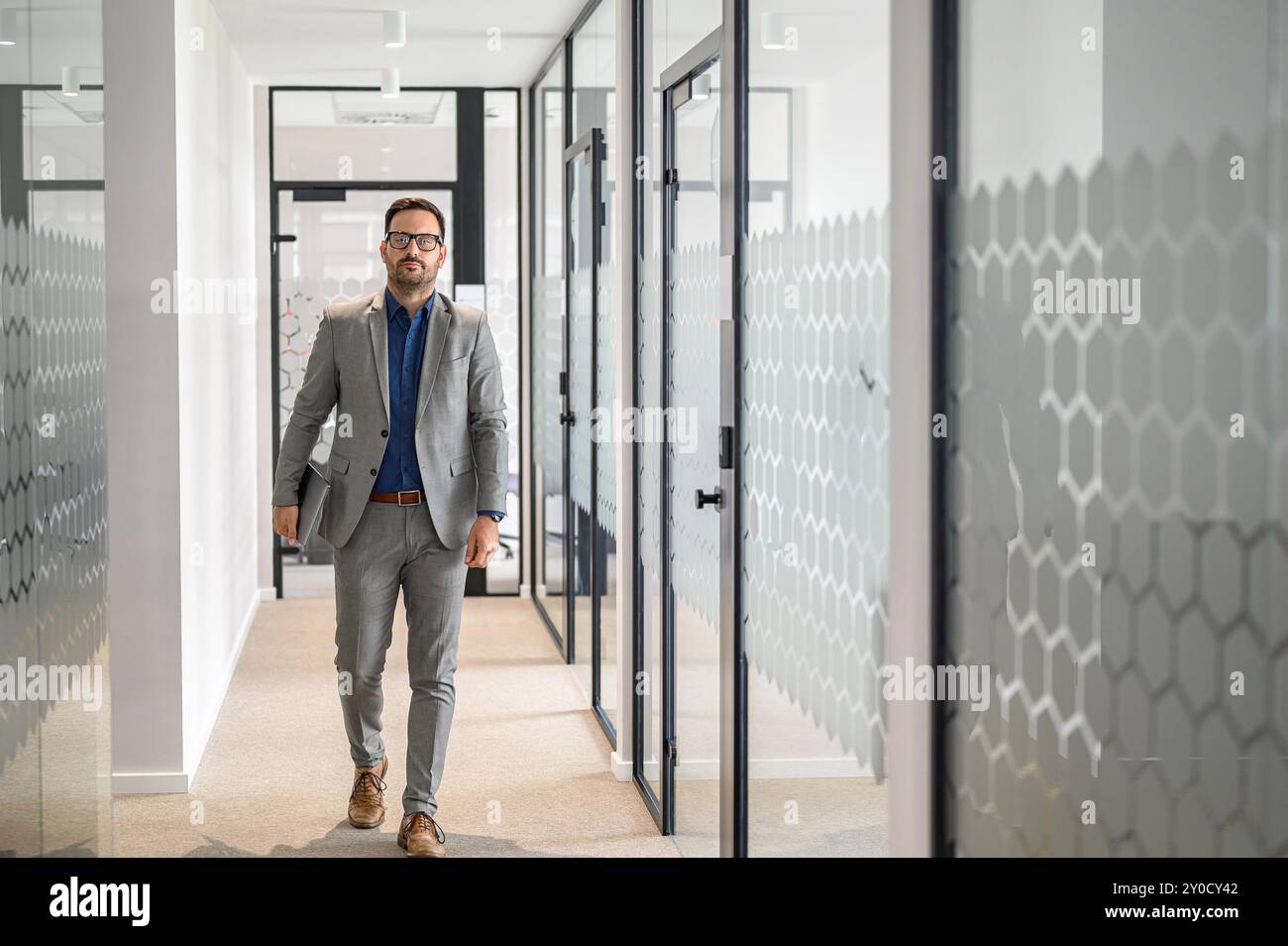 Portrait d'un gestionnaire masculin sérieux dans des lunettes tenant un ordinateur portable et marchant en toute confiance dans le couloir du bureau Banque D'Images