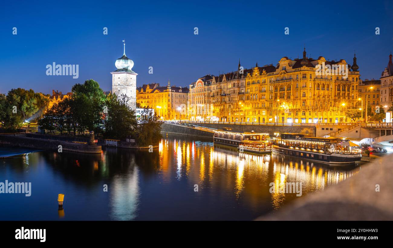 Le château d'eau de Sitkov se dresse le long du quai Masaryk à Prague, en Tchéquie, alors que le soleil se couche et que les lumières de la ville commencent à éclairer la scène. L'eau calme reflète la lueur chaude des bâtiments. Banque D'Images