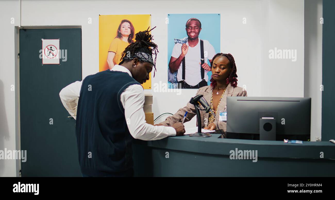 Employé de vente au détail ramassant les boîtes de commande de vêtements dans le grand magasin, signant la facture et les papiers de confirmation à la caisse enregistreuse. Femme afro-américaine donnant la marchandise demandée. Caméra B. Banque D'Images