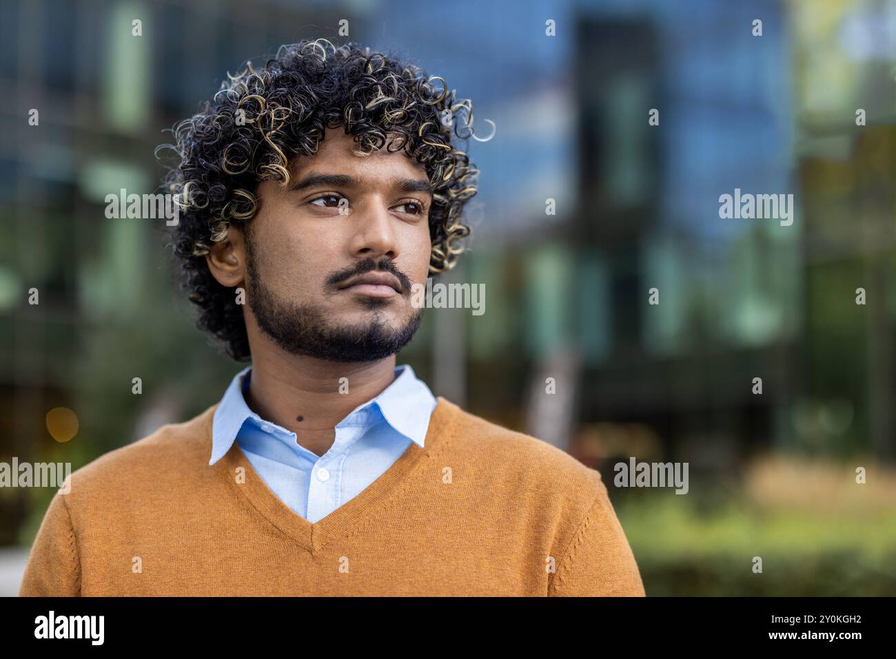 Jeune homme confiant avec des cheveux bouclés se tient à l'extérieur avec une expression réfléchie. Portant un pull orange décontracté et une chemise bleue, il respire le professionnalisme et la concentration. Banque D'Images