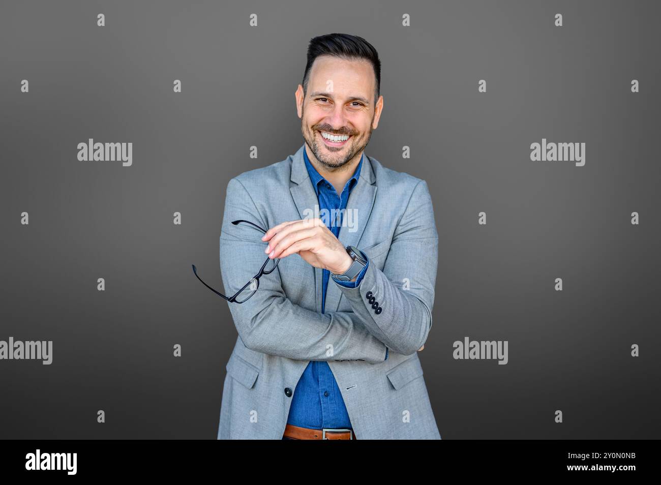 Portrait d'homme d'affaires réussi tenant des lunettes et avec le bras croisé posant sur fond gris Banque D'Images