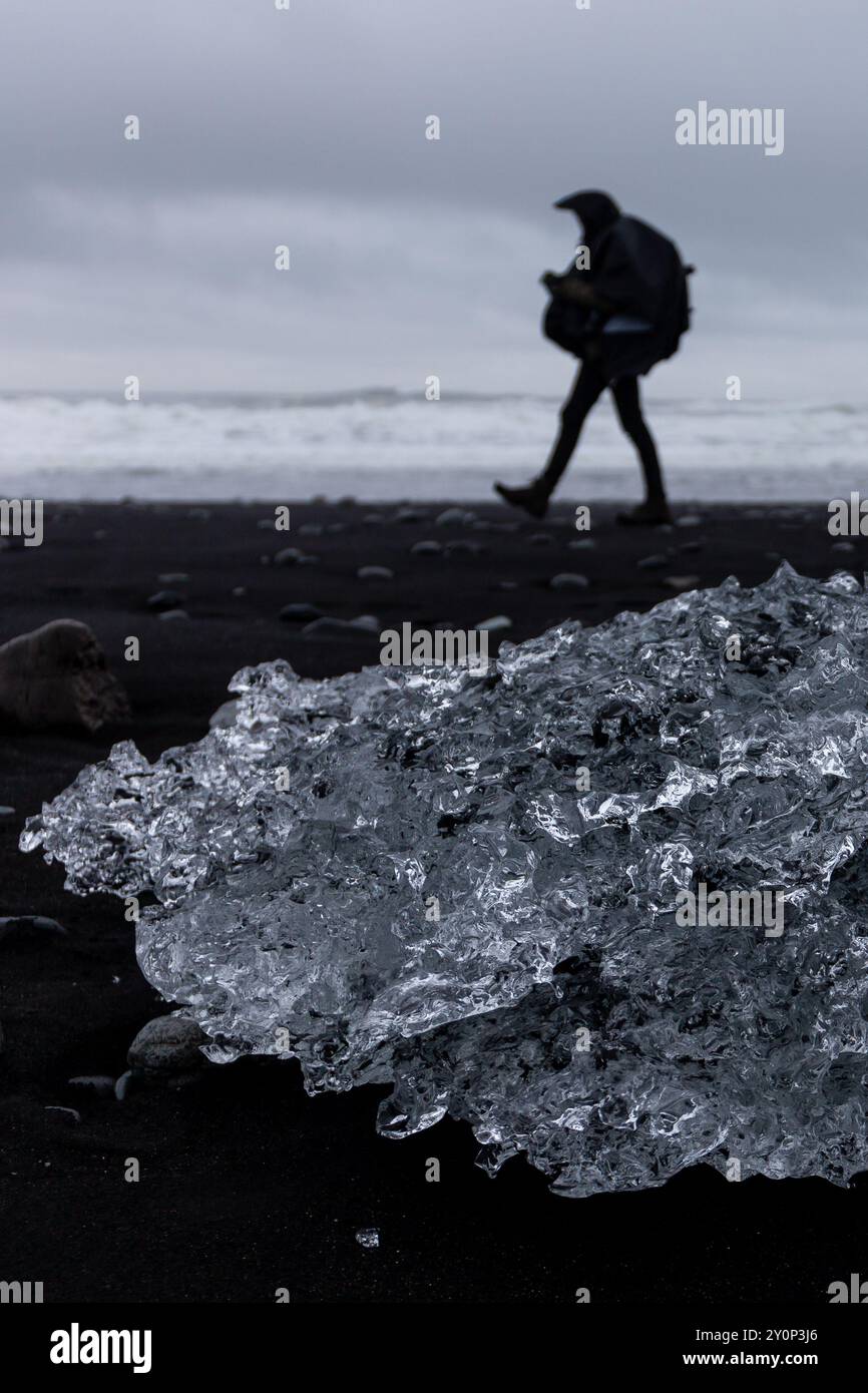 Iceberg transparent gisant sur du sable noir volcanique sur Diamond Beach en Islande, avec la silhouette d'une personne marchant avec un sac à dos. Banque D'Images