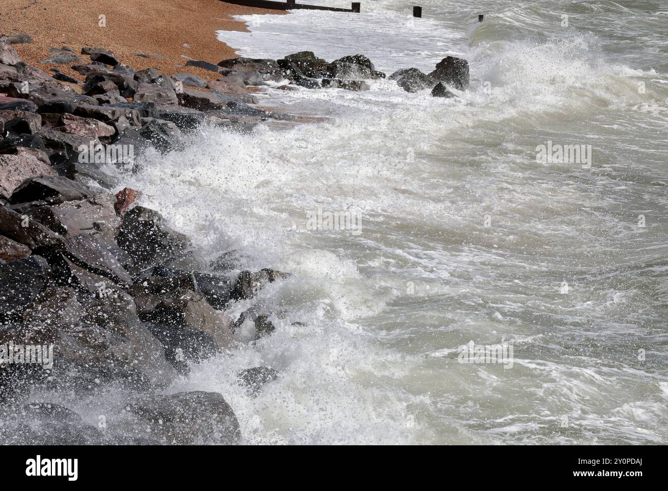 Vagues se brisant contre les rochers gris lors de conditions venteuses à la plage, sable et groynes en arrière-plan Banque D'Images