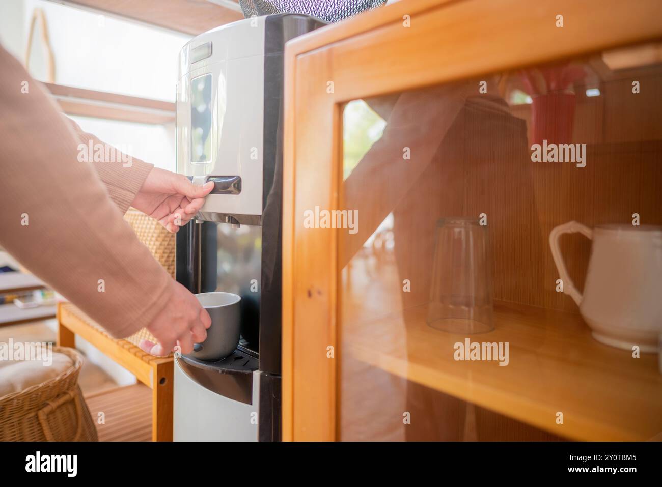 Une femme remplit une tasse à partir d'un distributeur de refroidisseur d'eau. Le distributeur de refroidisseur d'eau est sur une étagère en bois à côté d'une armoire en verre Banque D'Images