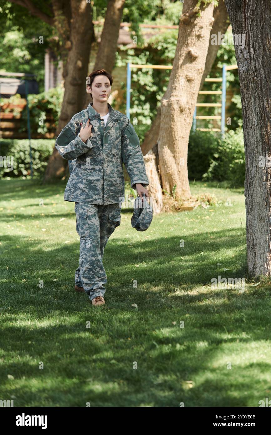 Un jeune soldat en uniforme de camouflage marche en toute confiance à travers un espace extérieur vert, embrassant le retour au pays. Banque D'Images