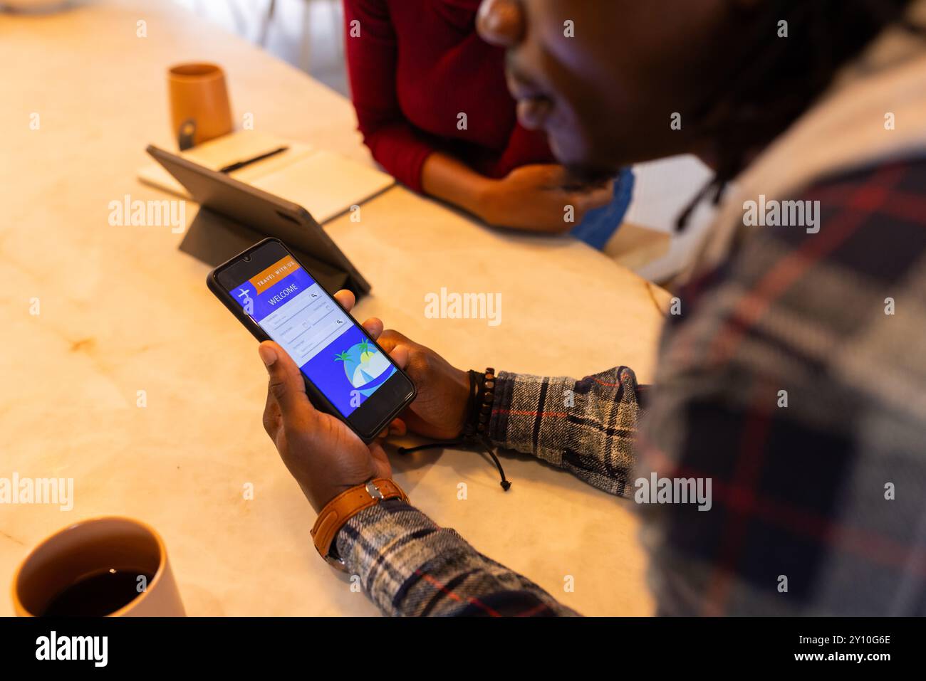 Tenant smartphone, homme afro-américain vérifiant l'application de réservation de vol à la maison avec tablette à proximité Banque D'Images