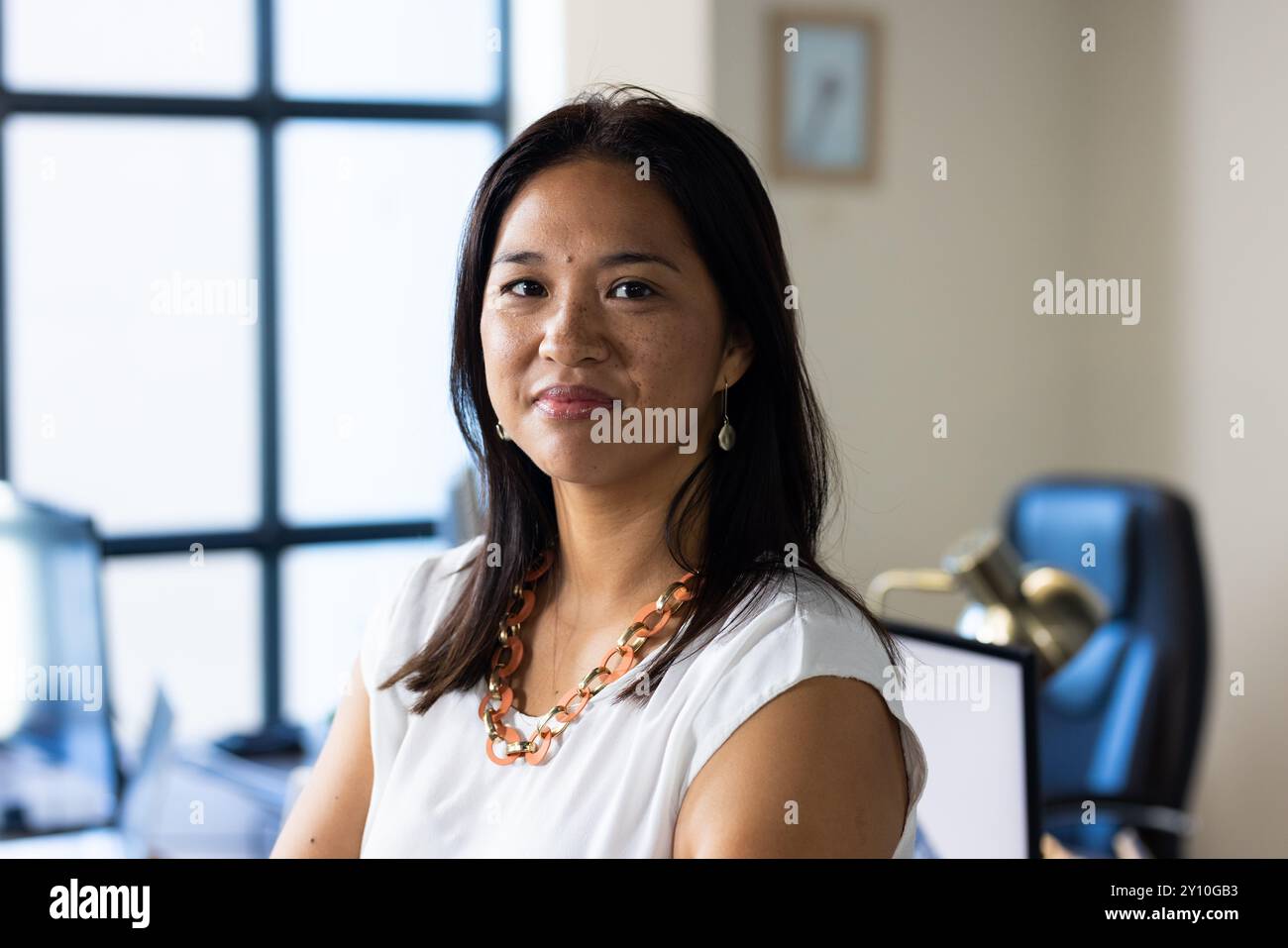 Femme souriante au bureau portant chemisier blanc et collier, debout avec confiance Banque D'Images