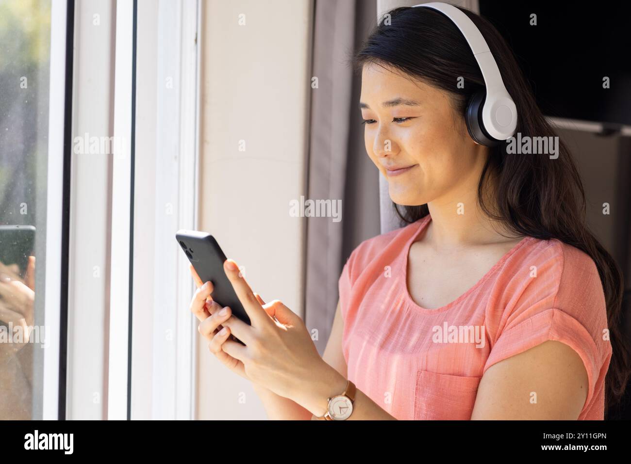 Écouter de la musique sur des écouteurs, femme asiatique utilisant un smartphone par fenêtre, souriant Banque D'Images