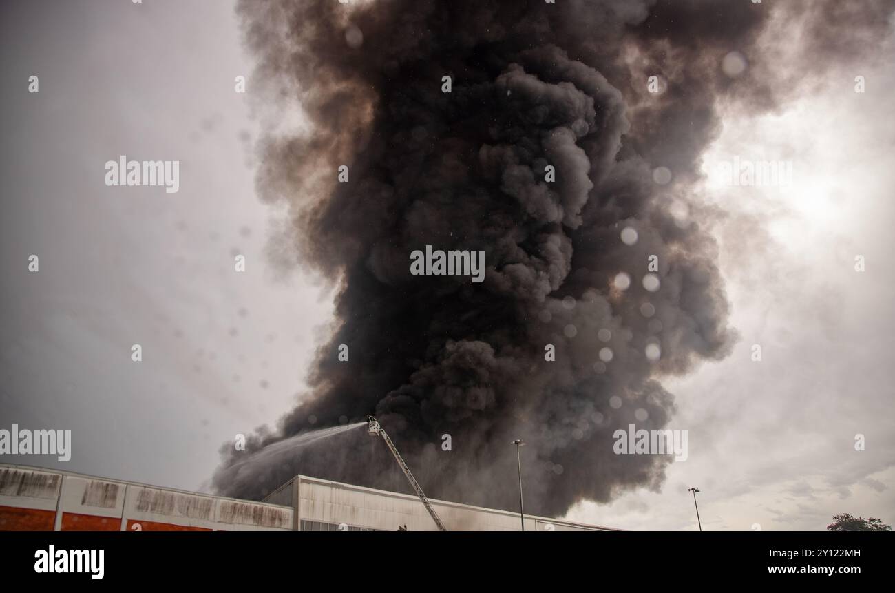 Un pompier pulvérise de l'eau à partir d'une échelle hydraulique pour éteindre un grand incendie industriel, avec une épaisse fumée noire qui jaillit dans le ciel Banque D'Images