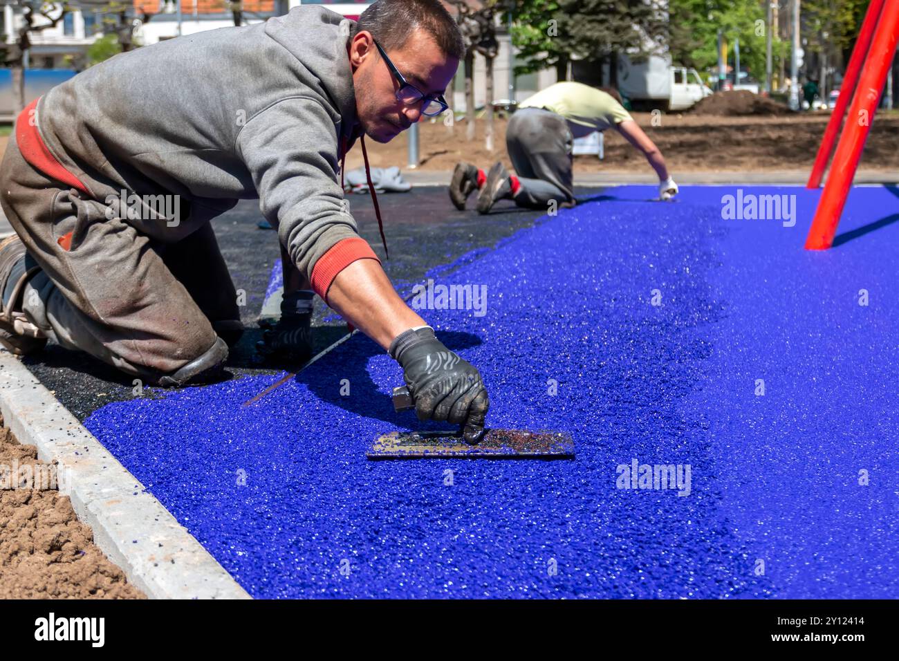 Belgrade, Serbie - 28 avril 2023 : Nouvelle aire de jeux pour enfants en construction. Une main de maçon avec l'étalement de truelle et nivellement de miettes de caoutchouc mou RU Banque D'Images