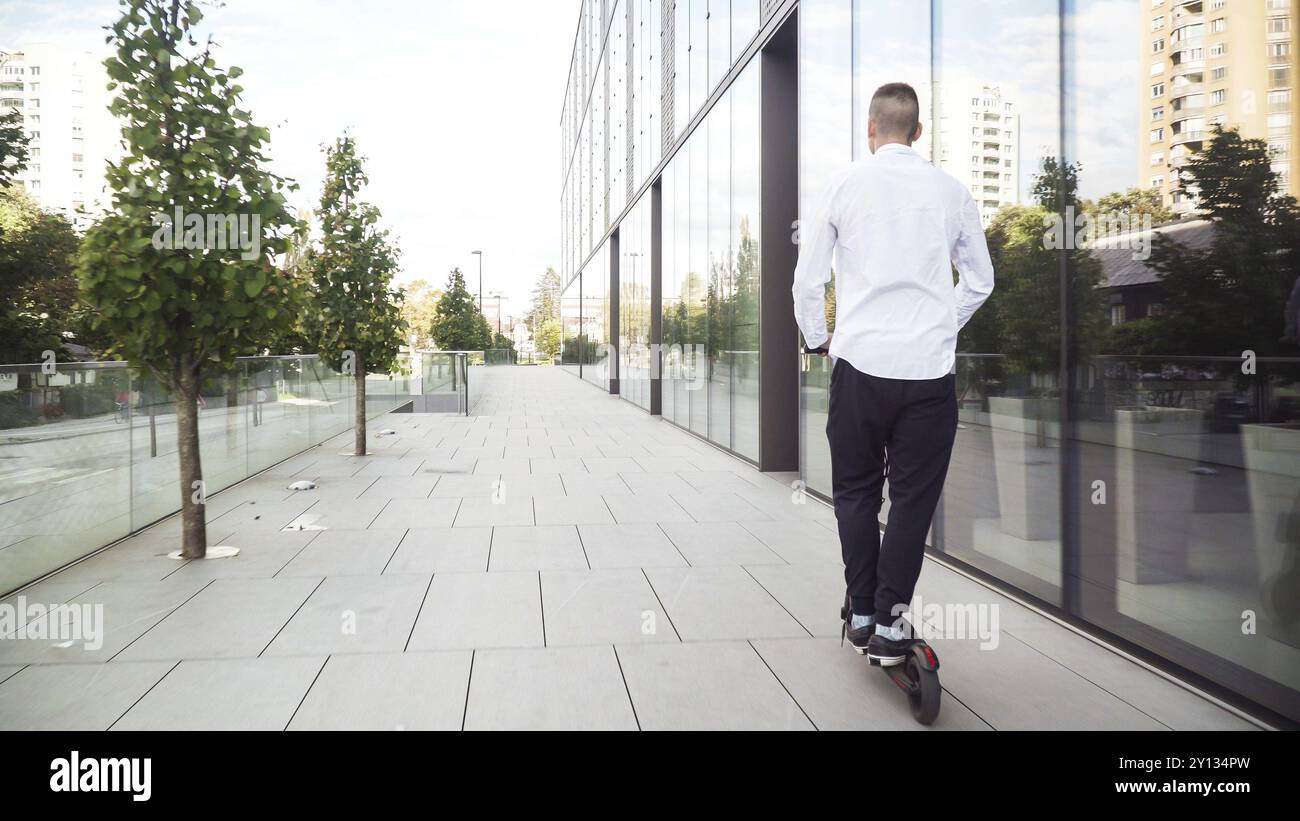 Jeune homme d'affaires décontracté qui monte en scooter électrique sur un trottoir dans une ville d'affaires Banque D'Images