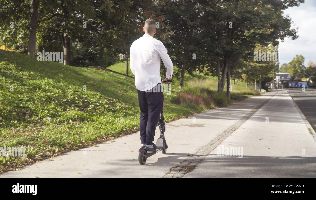 Jeune homme d'affaires décontracté qui monte en scooter électrique sur un trottoir en été Banque D'Images