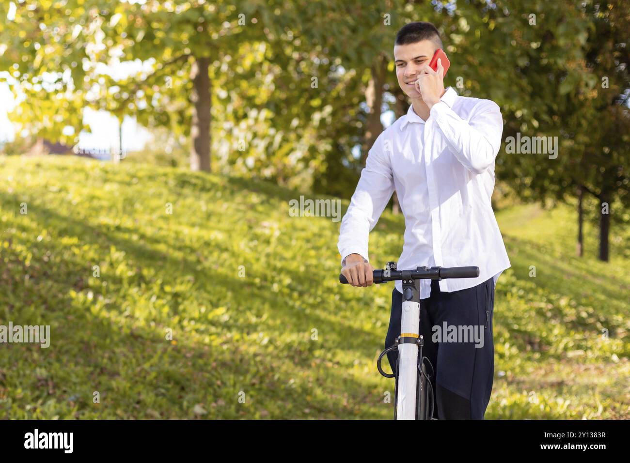 Joyeux et casual man riding scooter électrique dans le parc de la ville et parlant au téléphone Banque D'Images