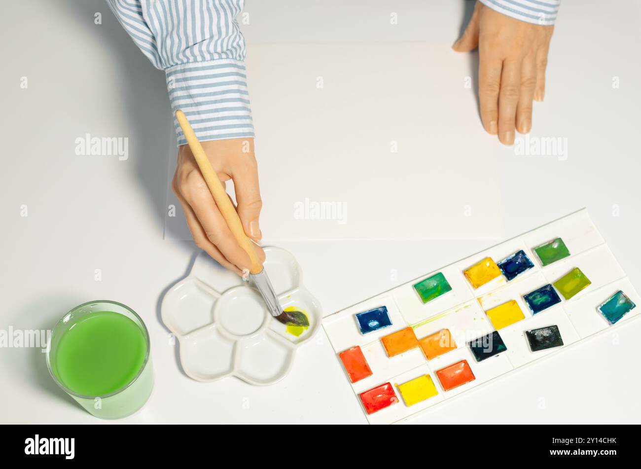Artiste mélangeant des peintures aquarelles dans la palette céramique en studio, gros plan. Mains féminines et accessoires d'aquarelle sur la table, vue de dessus Banque D'Images