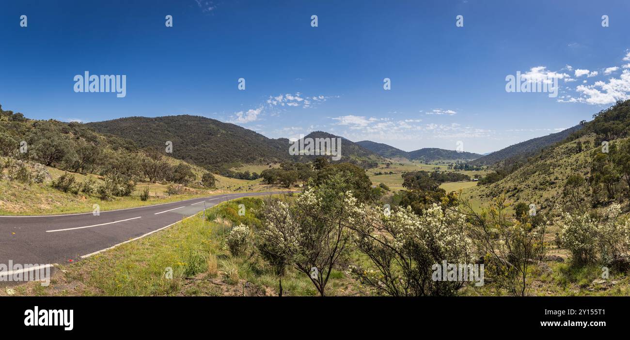 Ce paysage présente une route sinueuse flanquée de verdure luxuriante, de collines ondulantes et de vastes champs ouverts, le tout illuminé par un soleil chaud et parsemé de cl Banque D'Images