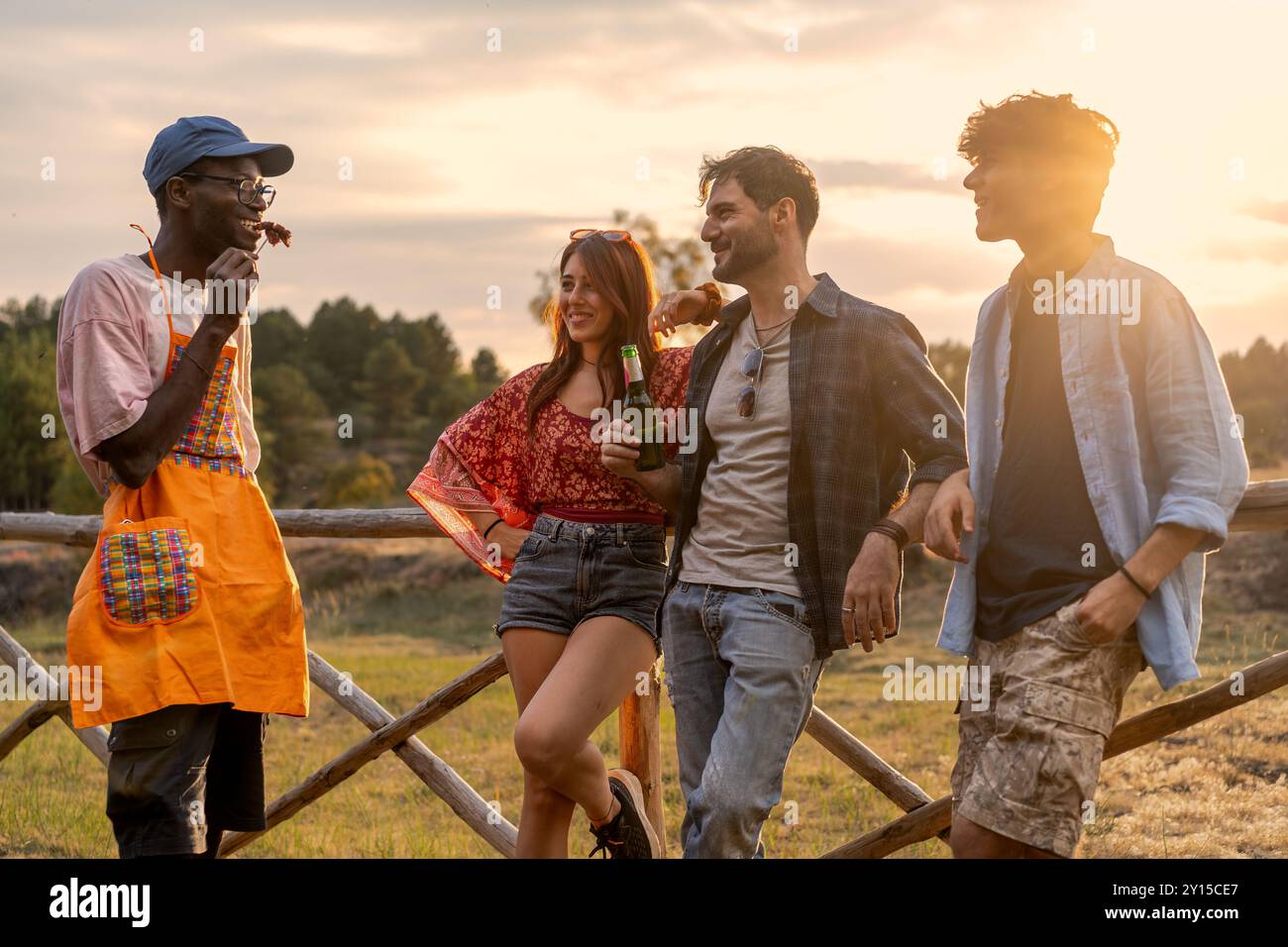 Groupe de jeunes adultes diversifiés socialisant à l'extérieur au coucher du soleil. Amis multiculturels partageant des boissons et des rires dans un cadre rural, mettant en valeur inclusi Banque D'Images