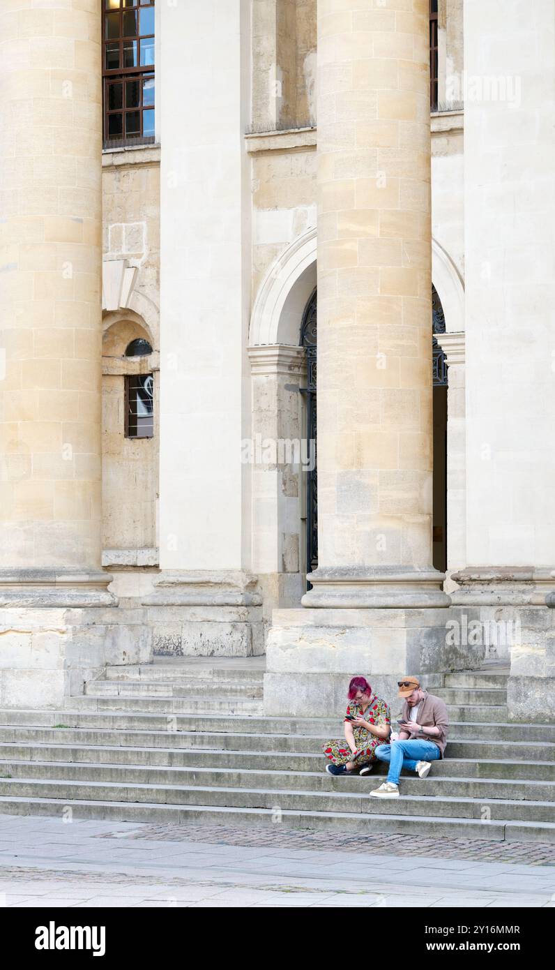 Quelques visiteurs sont assis sur les marches à l'extérieur de l'ancienne bibliothèque, qui fait partie de la bibliothèque Bodleian de l'Université d'Oxford, en Angleterre. Banque D'Images