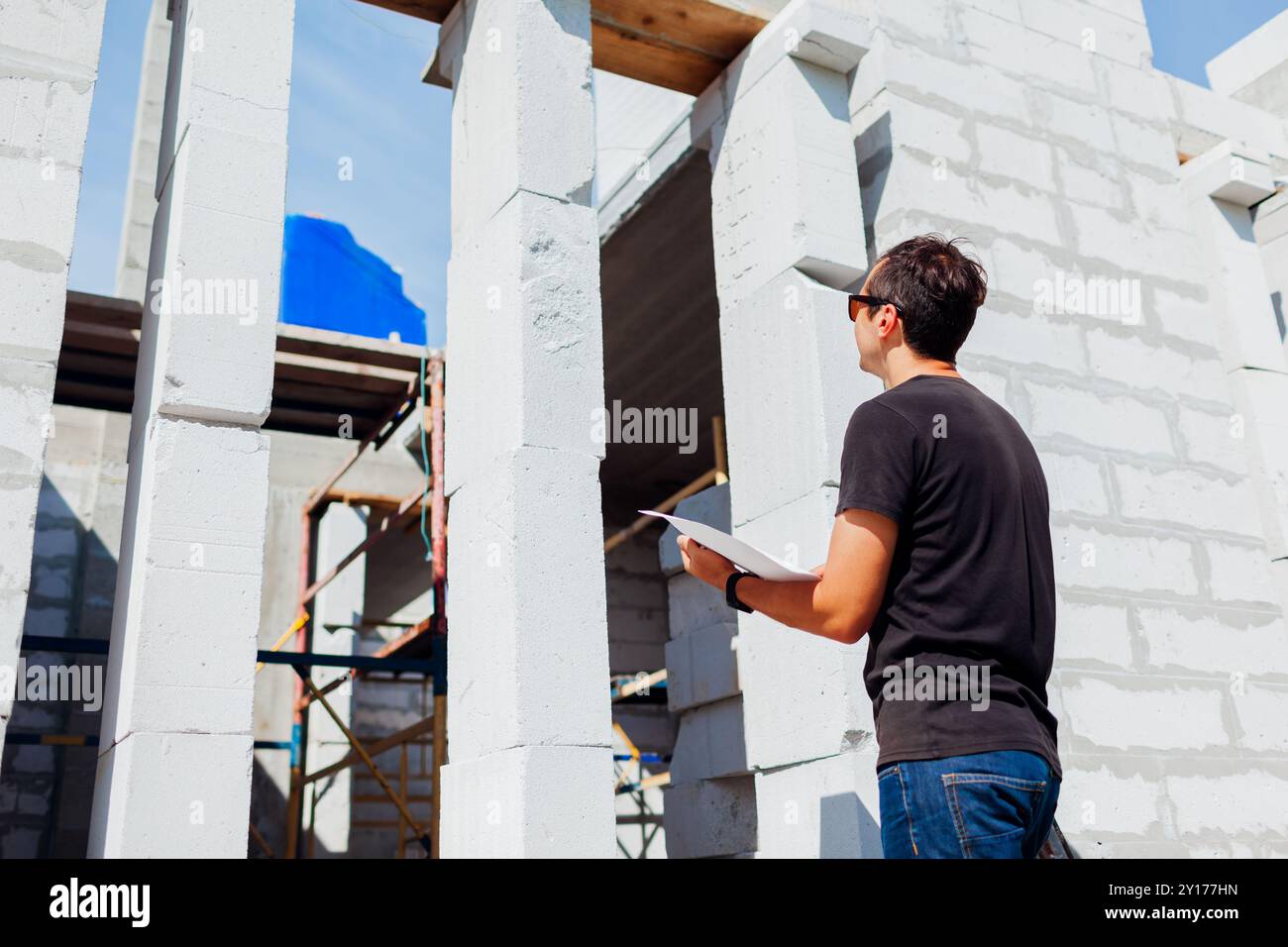 Homme architecte ingénieur vérifiant la maison en construction comparant avec le plan de plan sur papier. Le travailleur regarde la fente de la fenêtre à l'extérieur Banque D'Images