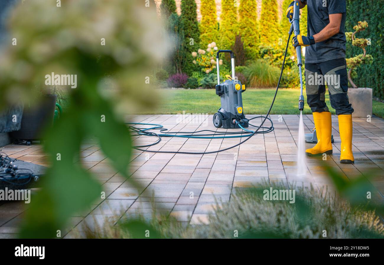 Un ouvrier en bottes jaunes utilise une laveuse haute pression pour nettoyer les carreaux de patio, entouré de verdure luxuriante dans un beau jardin. Banque D'Images