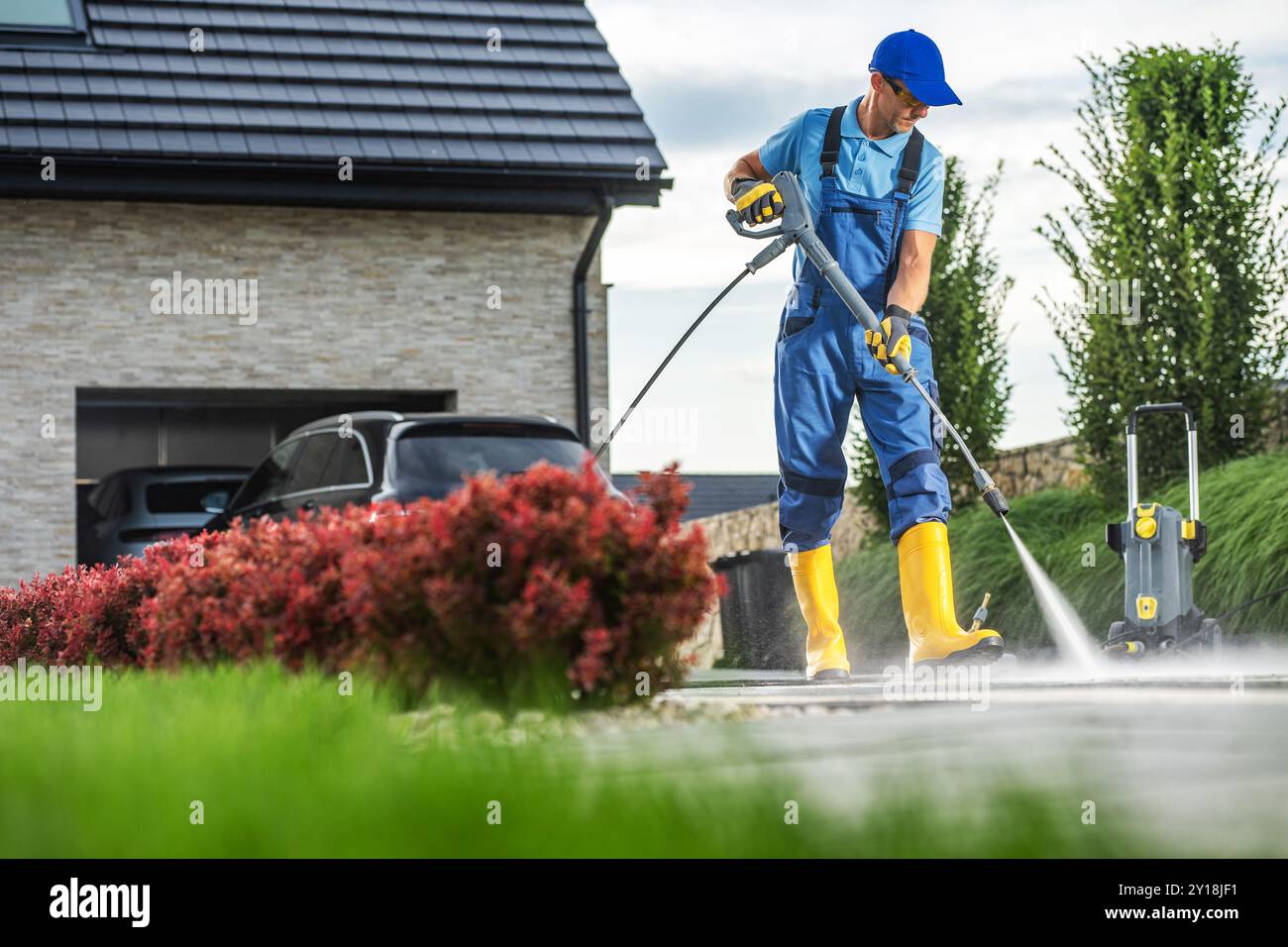 Un homme en salopette bleue et bottes jaunes nettoie une allée avec un nettoyeur haute pression, entouré de verdure luxuriante et de fleurs vibrantes par un après-midi lumineux Banque D'Images