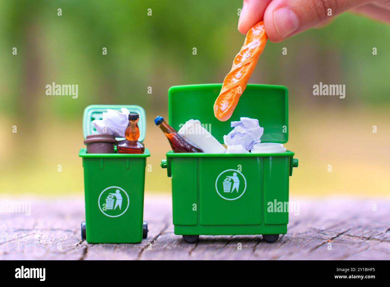 Gros plan d'une main plaçant une baguette dans une poubelle verte miniature avec des ordures non triées, mettant en évidence des concepts inappropriés d'élimination des déchets et de recyclage. Banque D'Images