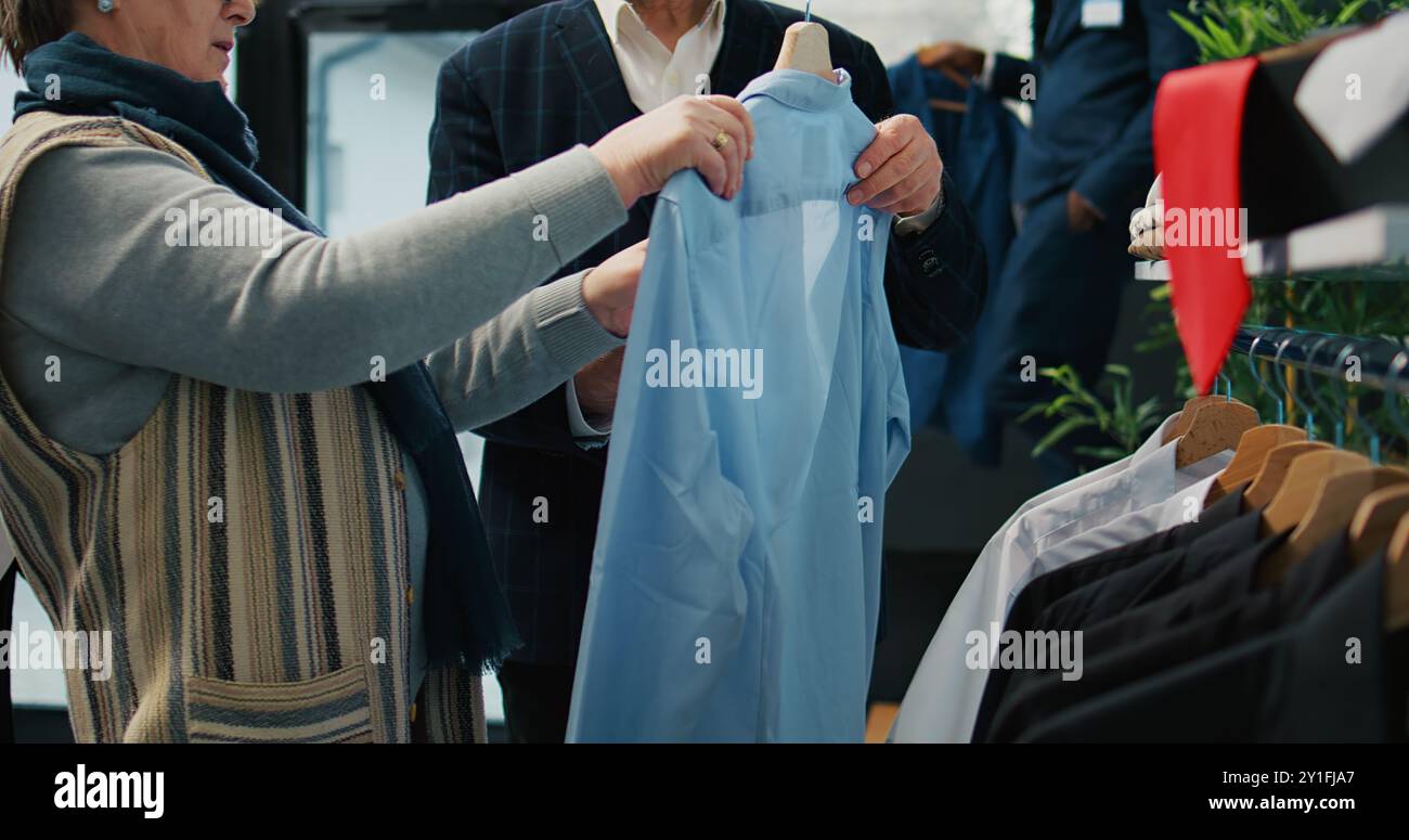 Femme à la recherche de la bonne chemise formelle pour son mari, cherchant à élargir la garde-robe des ventes à la boutique de mode. Couple senior shopping pour costumes et articles élégants, activité commerciale. Caméra B. Banque D'Images