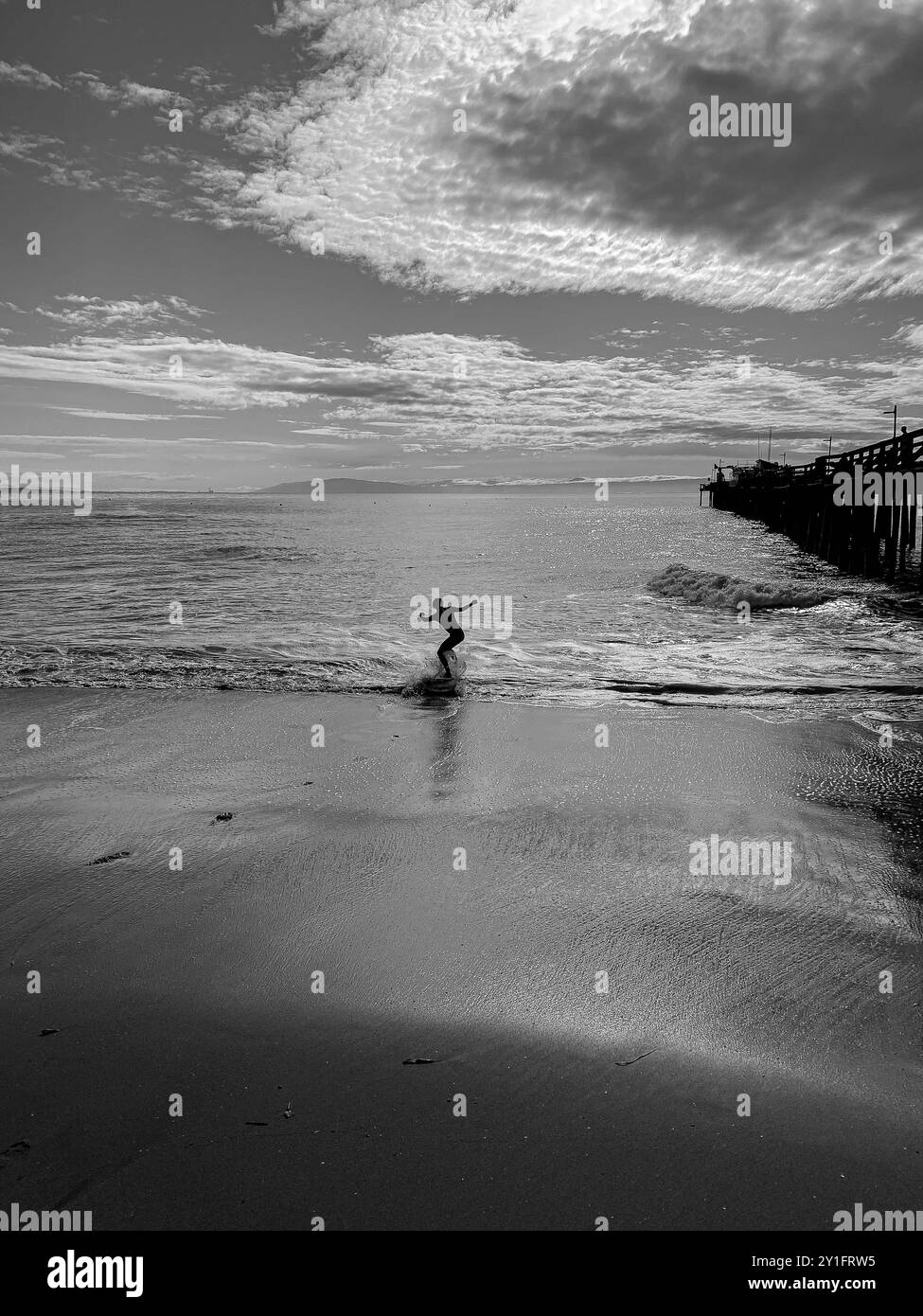 Un skimboarder glisse le long des eaux peu profondes de Capitola Beach, en Californie, alors que les vagues s'écrasent près de la jetée sous un ciel spectaculaire et rempli de nuages à Thi Banque D'Images