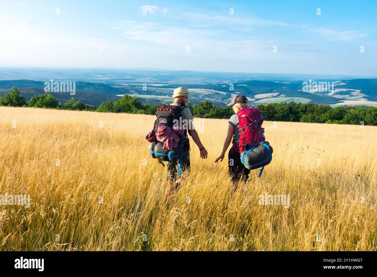 Deux randonneurs avec sacs à dos trekking, un couple marchant à travers une prairie herbeuse dorée vers une vue lointaine de collines ondulantes et un ciel bleu clair Banque D'Images