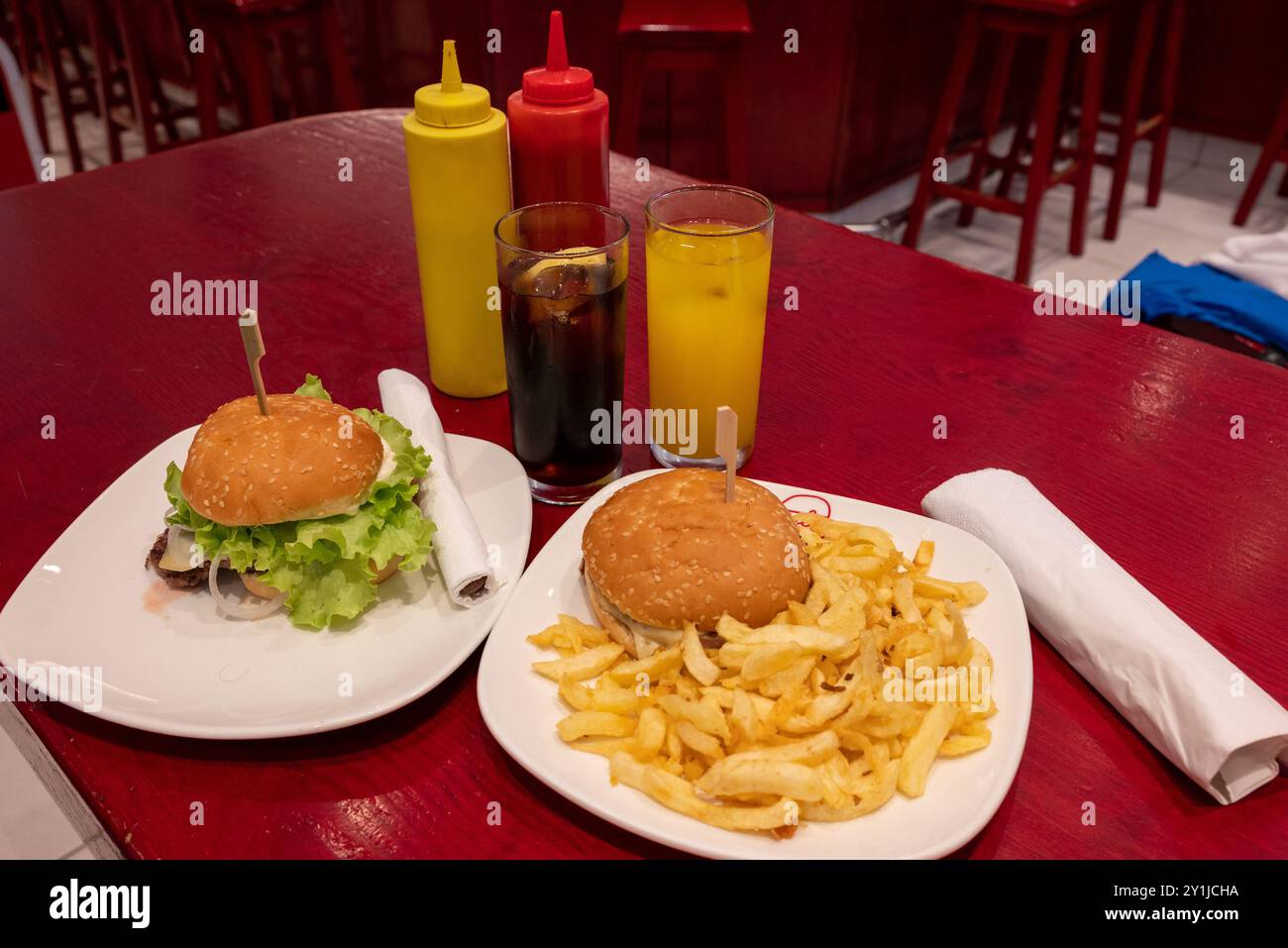 Repas pour deux dans un fast-food : deux savoureux burgers grillés avec frites, soda et jus d'orange sur plats blancs et table en bois rouge, avec dis en plastique Banque D'Images