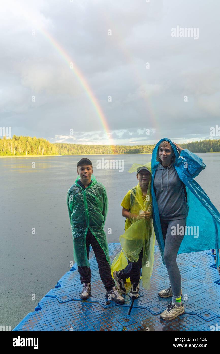 Famille dans les imperméables profiter d'une vue sur le lac avec un arc-en-ciel vif en arrière-plan. Le lac serein et les arbres luxuriants créent une atmosphère joyeuse sur un nuage Banque D'Images