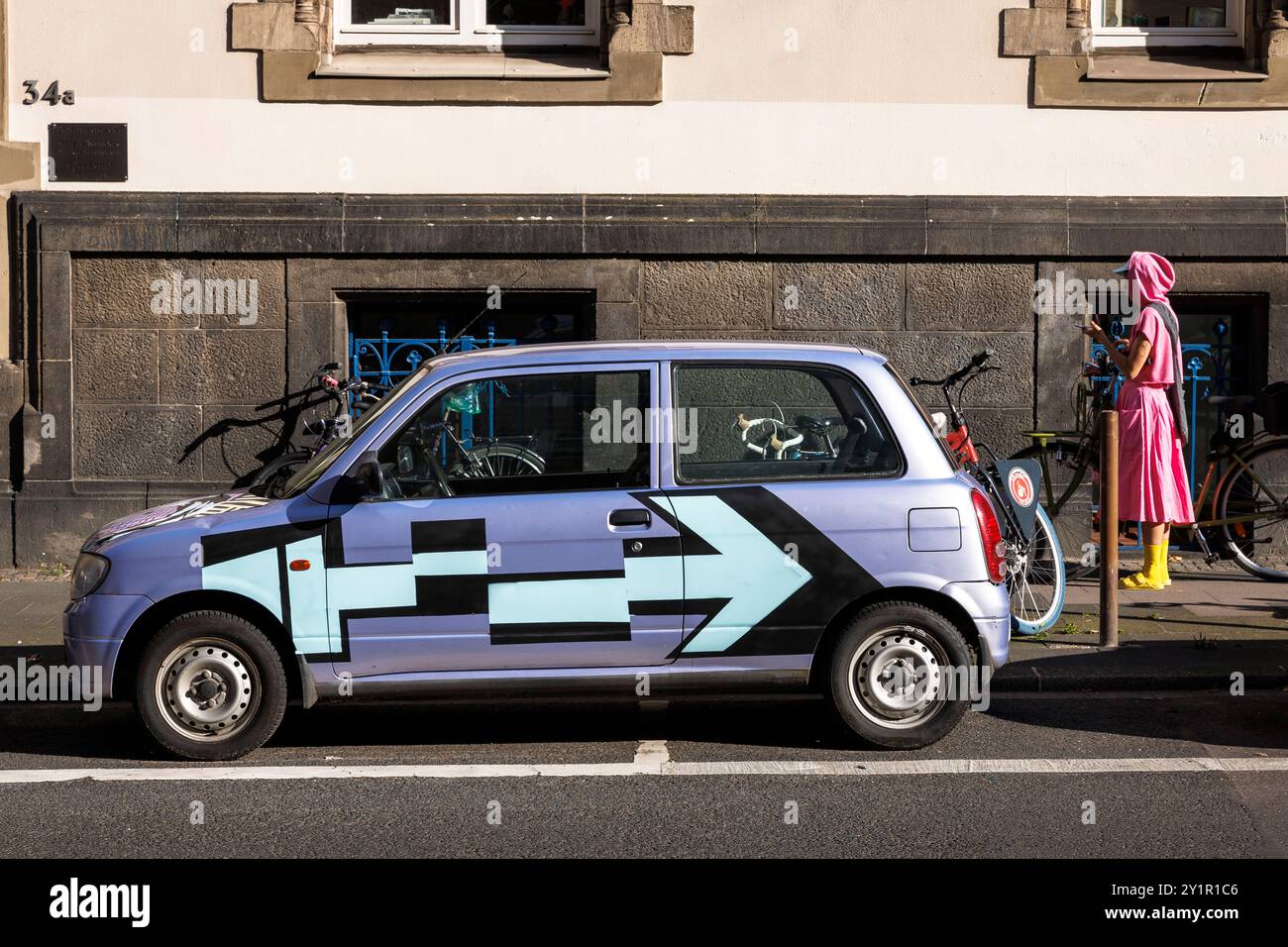 Femme en vêtements roses et jaunes debout à côté d'une petite voiture peinte flashy, Cologne, Allemagne. Frau in rosa und gelber Kleidung steht neben einem auf Banque D'Images