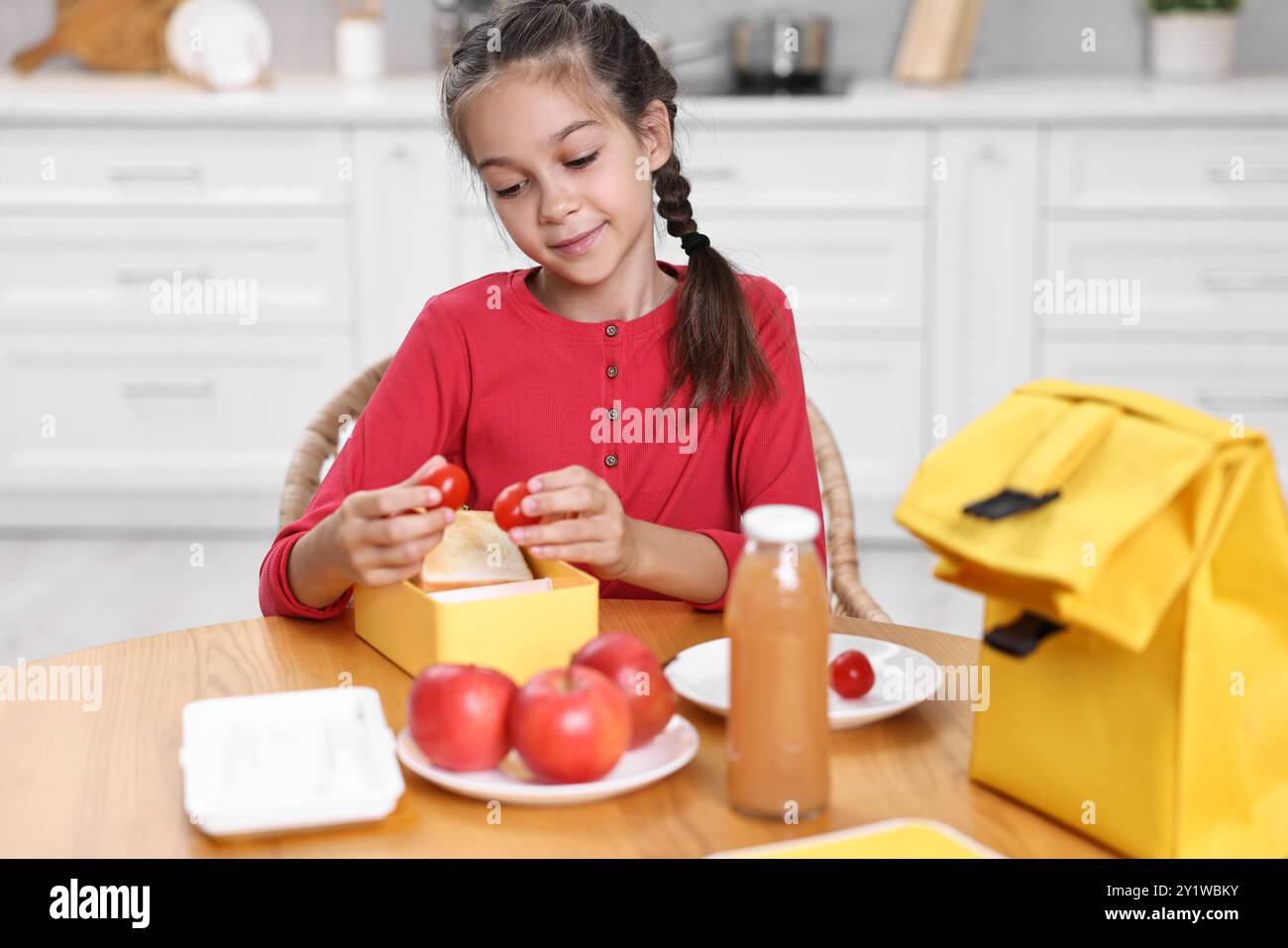 Fille mignonne mettant des tomates dans la boîte à lunch à la table en bois dans la cuisine. Préparation à l'école Banque D'Images