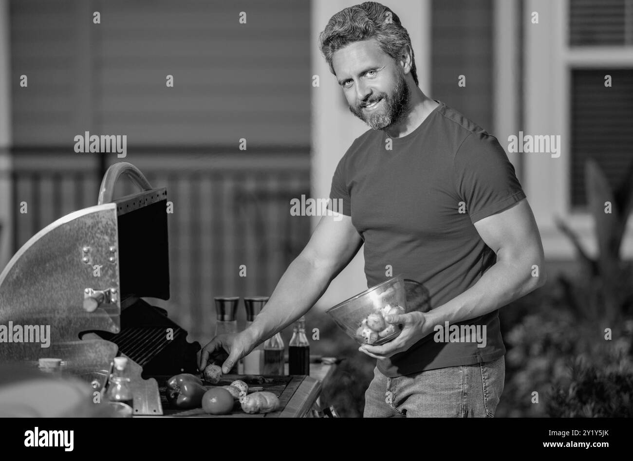 Le barbecue. Homme d'âge moyen en t-shirt pour barbecue. Rôtir et griller des aliments. Homme tenir des ustensiles de cuisine barbecue. Rôtir de la viande à l'extérieur Banque D'Images