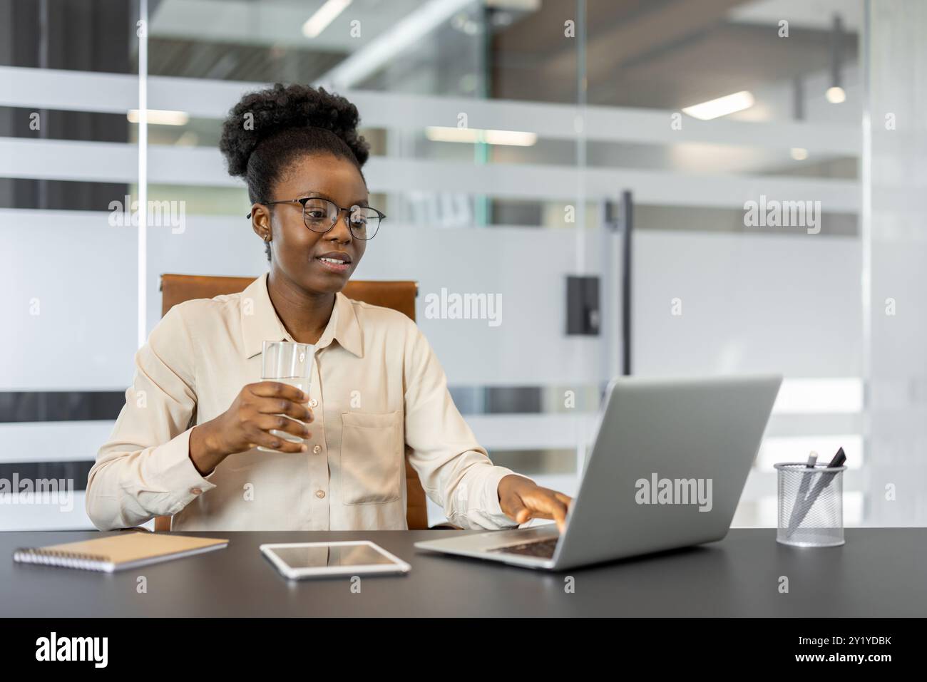 Femme d'affaires afro-américaine confiante utilisant un ordinateur portable dans un cadre de bureau. Elle tient un verre d'eau, avec tablette et téléphone visibles sur le bureau. L'atmosphère professionnelle suggère la concentration Banque D'Images
