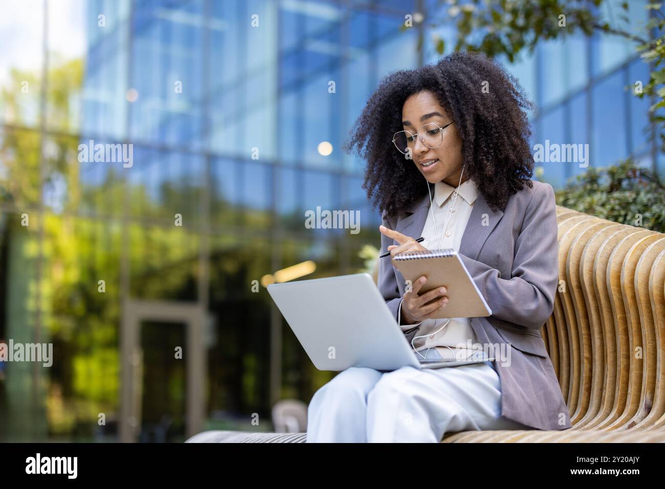 Femme d'affaires afro-américaine participe à un cours vidéo en ligne à l'extérieur. Elle communique avec l'instructeur via un ordinateur portable, prend des notes dans les utilisations de l'ordinateur portable pour les appels vidéo, améliorant ainsi ses compétences professionnelles Banque D'Images