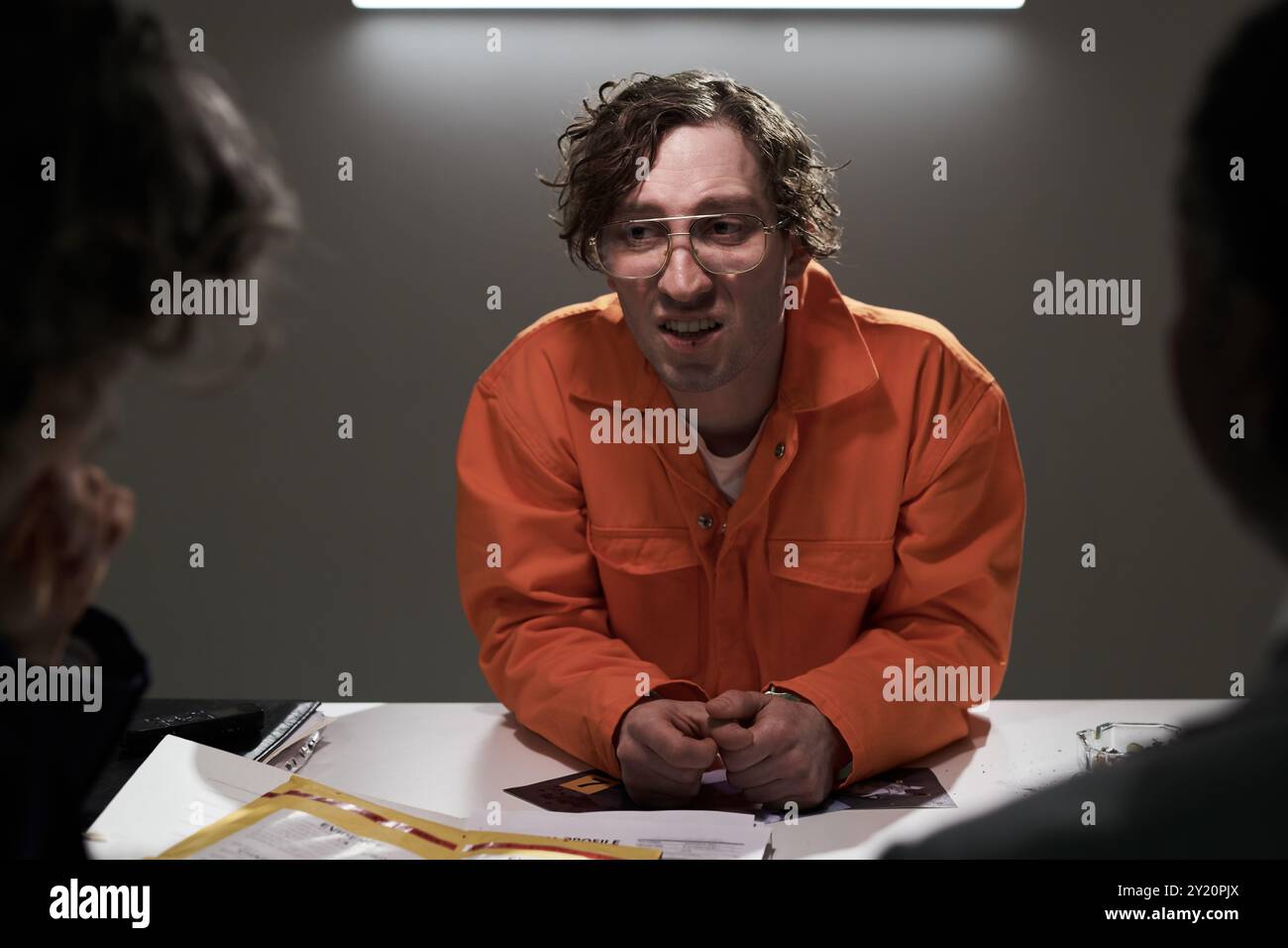 Homme en combinaison orange assis à table dans la salle d'interrogatoire, peut-être engagé dans une conversation intense, avec d'autres en arrière-plan et des documents sur la table Banque D'Images