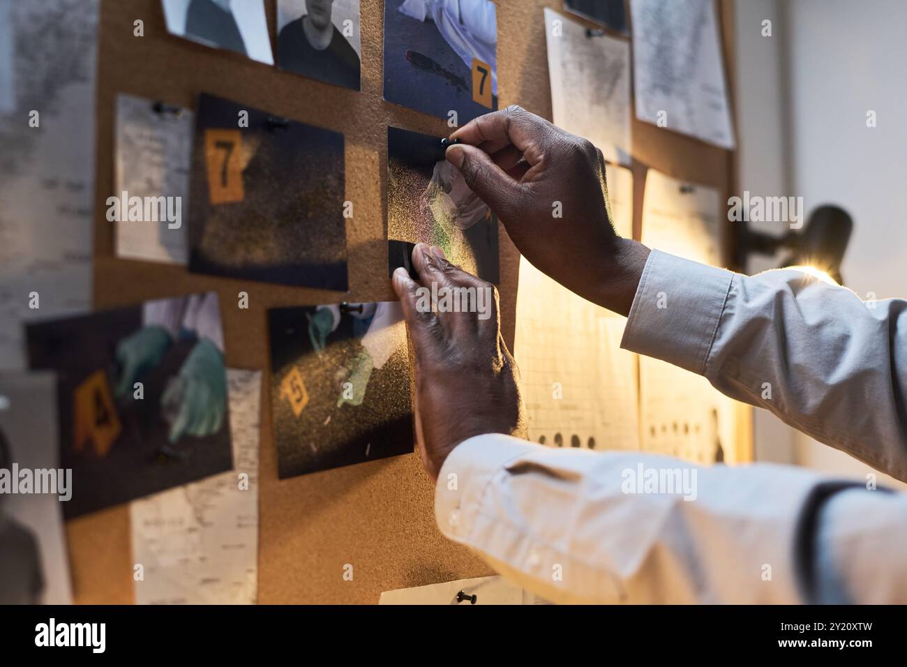 Mains épinglant des photos et des notes sur le tableau d'affichage dans une pièce faiblement éclairée, avec divers éléments de preuve liés à la scène de crime sous enquête Banque D'Images